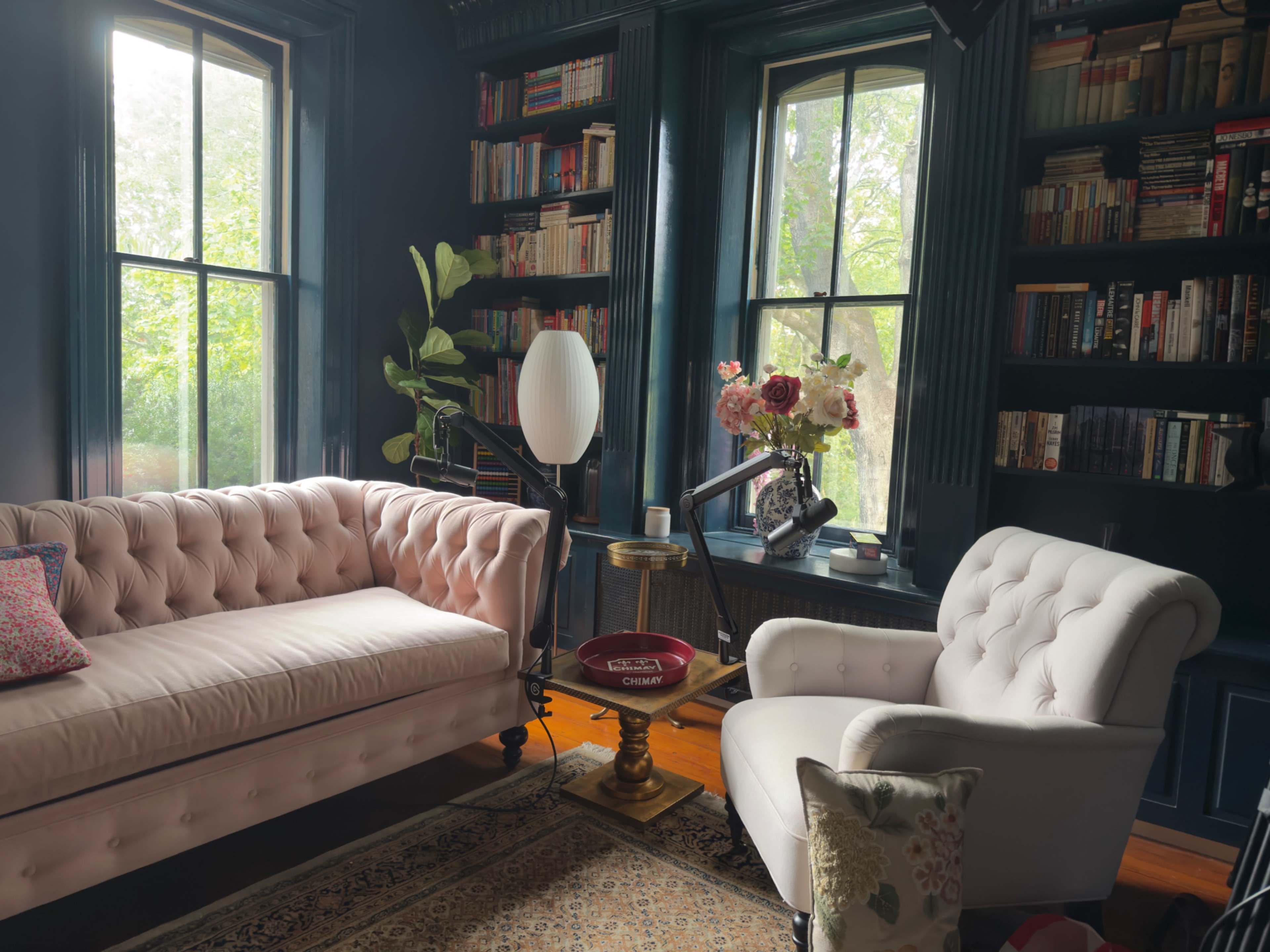 The image depicts a cozy reading nook featuring a pink tufted sofa and a white upholstered armchair, surrounded by bookshelves filled with books and a decorative flower arrangement.