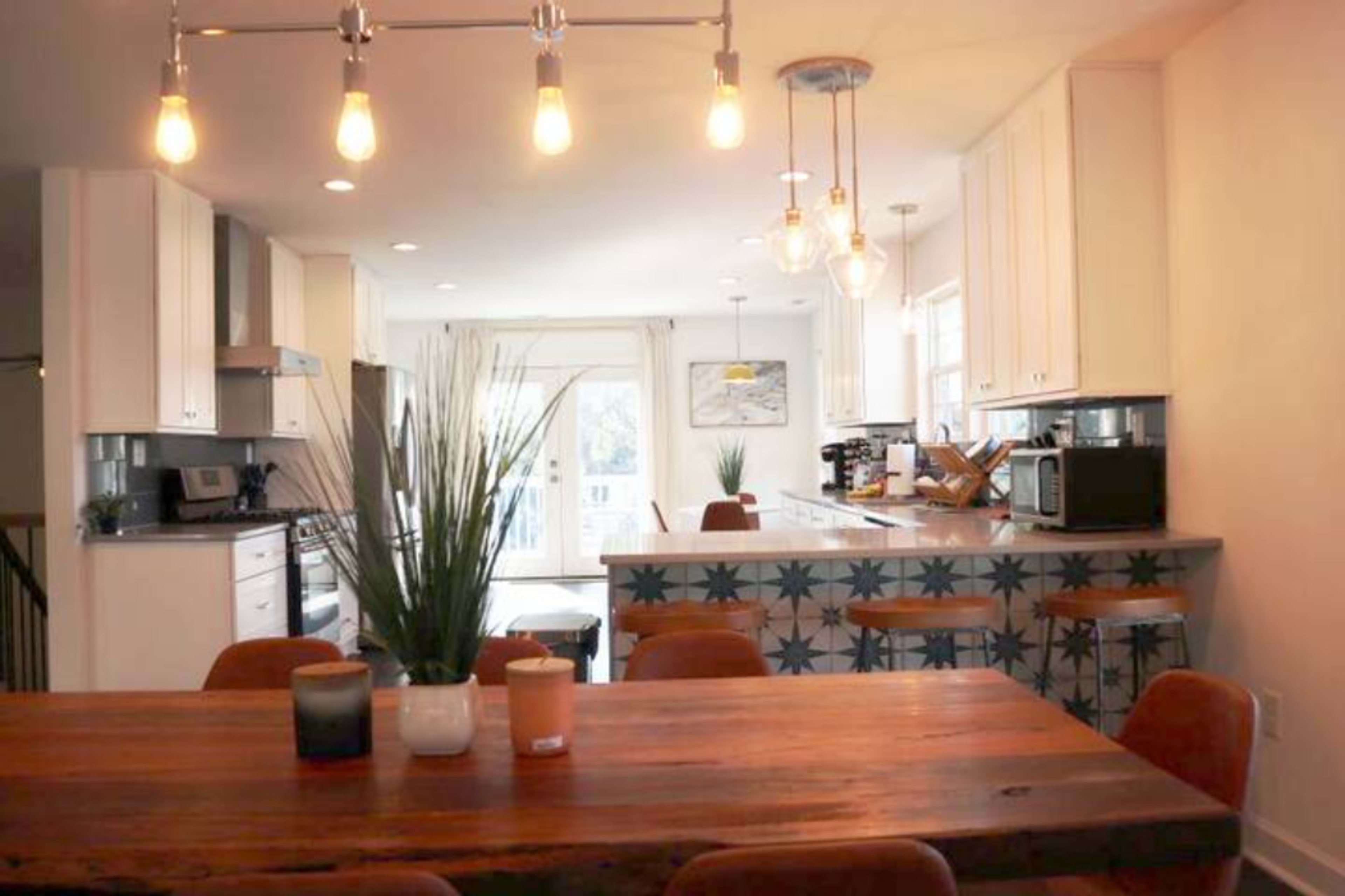The image shows a modern kitchen with a dining table, featuring wooden surfaces and a well-lit atmosphere, complemented by minimalistic decor and bar stools at the counter.