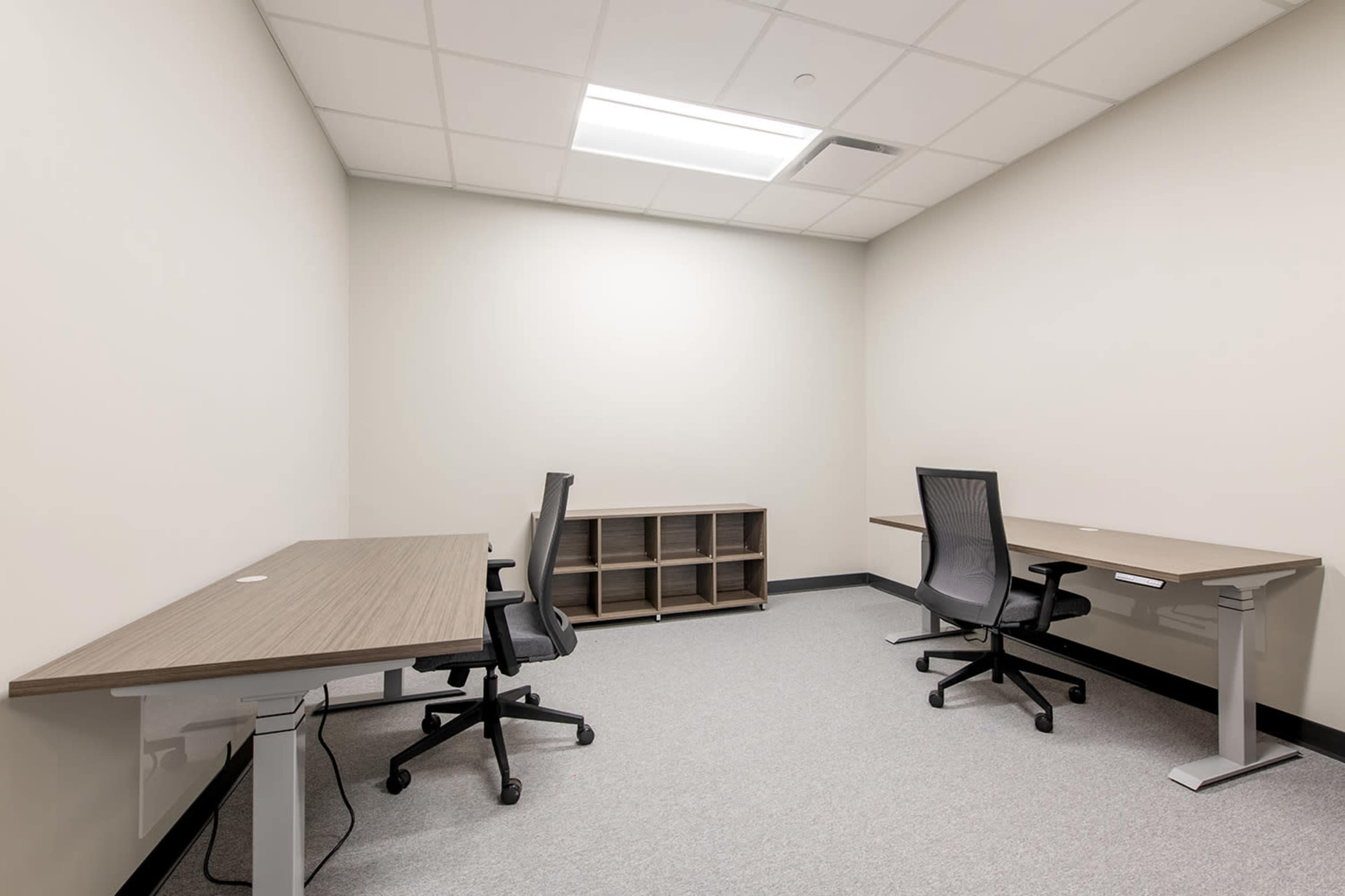 The image shows a sparsely furnished office space with two desks, two office chairs, and a small shelving unit along one wall.