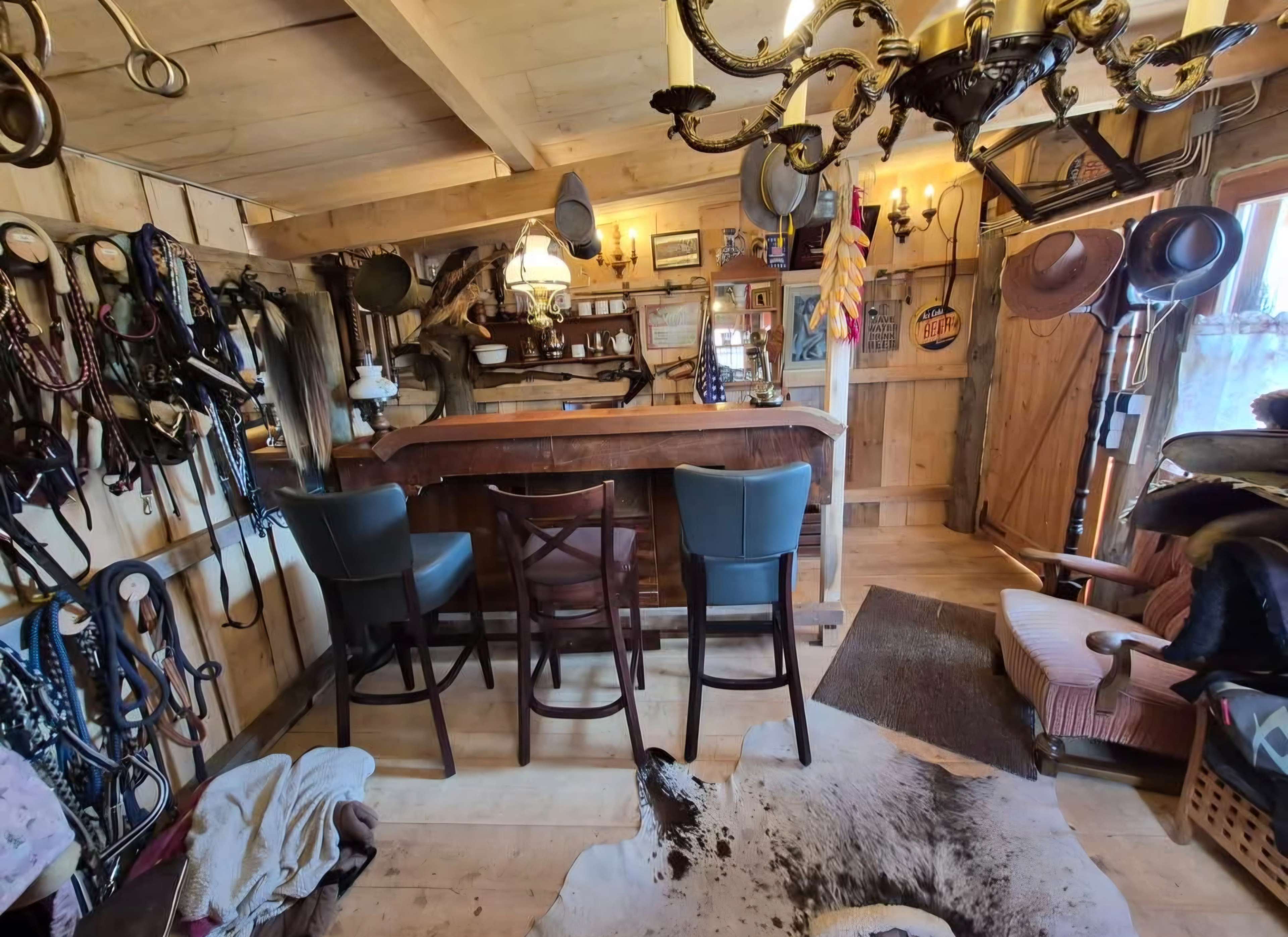 The image shows a rustic interior of a barn or lodge with a wooden bar, high chairs, and various horse riding gear hanging on the walls.