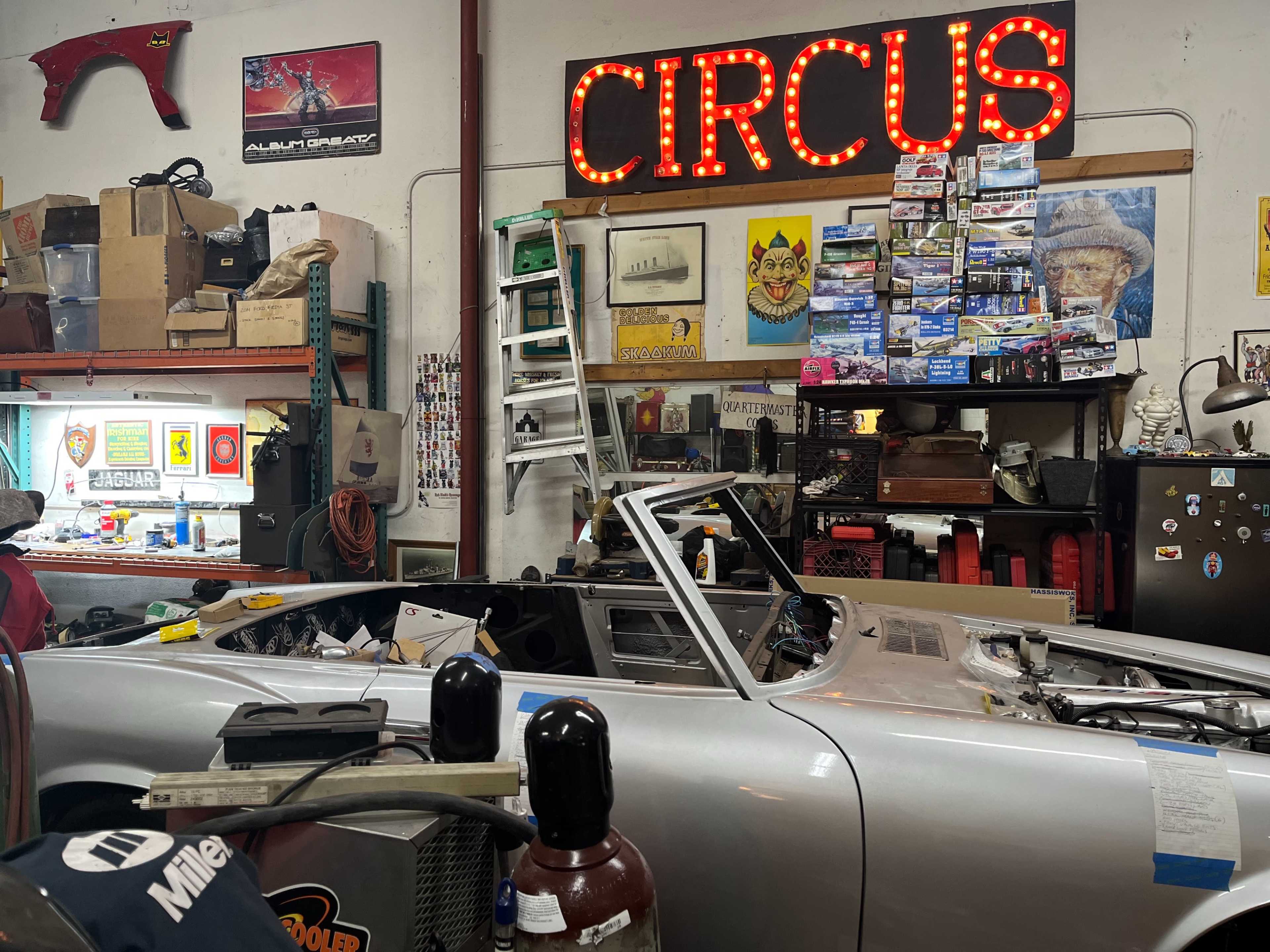 A silver convertible car is partially disassembled in a cluttered garage adorned with circus-themed decorations and various tools.