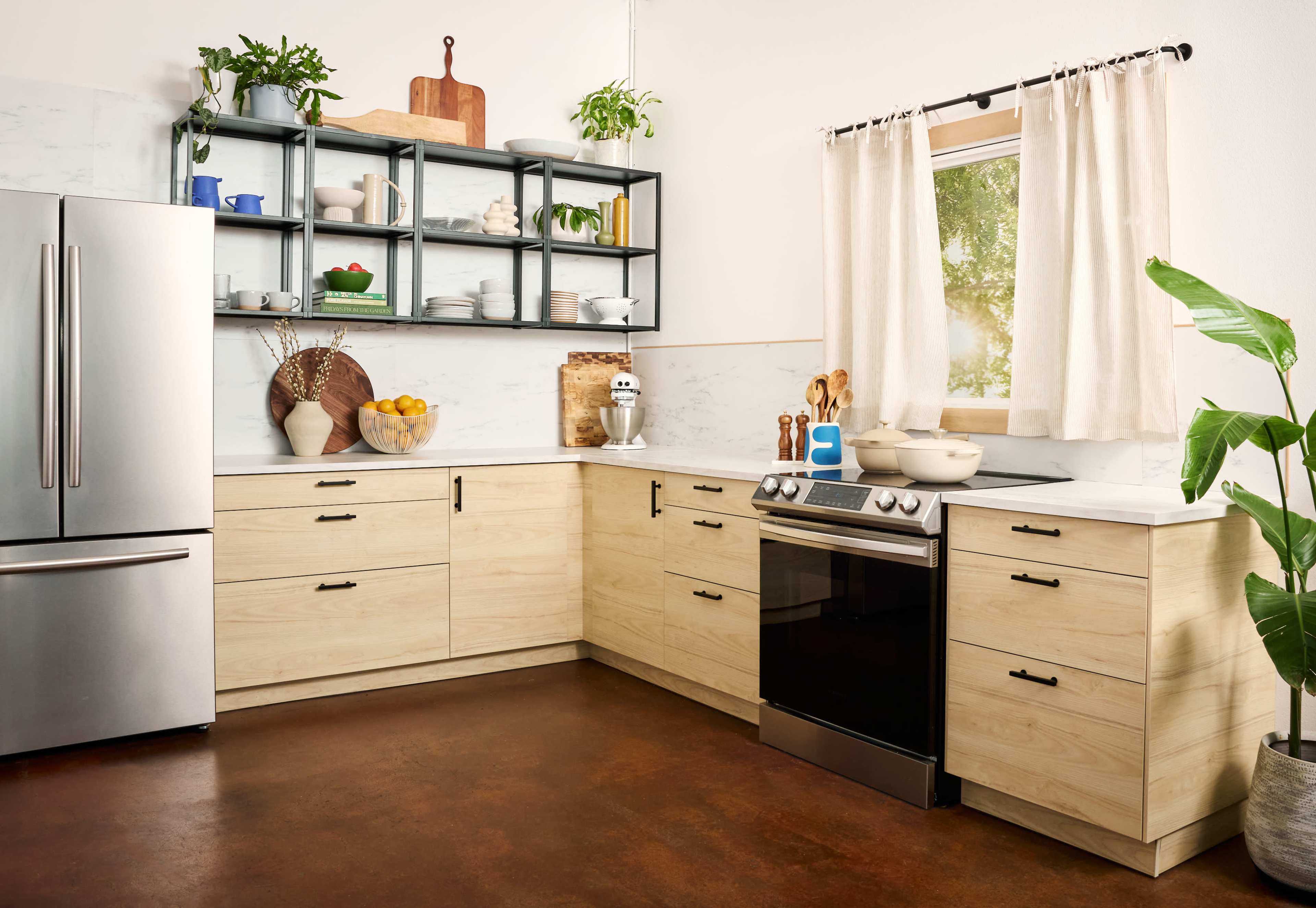 The image shows a modern kitchen with light wood cabinetry, a stainless steel refrigerator, an oven, and open shelving displaying plates and bowls.