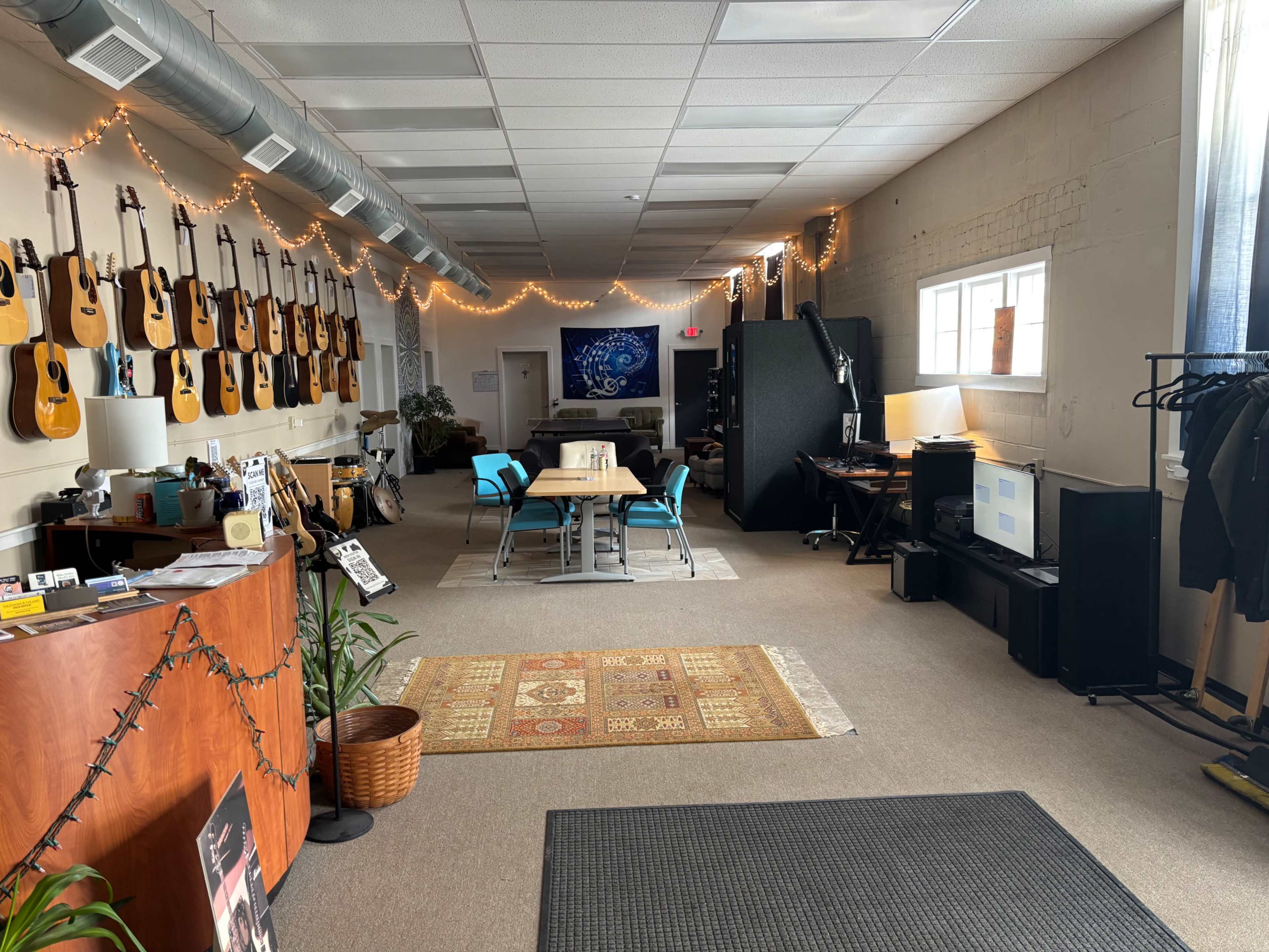 The image shows a spacious room with acoustic guitars displayed on the walls, a central table surrounded by blue chairs, and a reception area featuring a wooden desk.