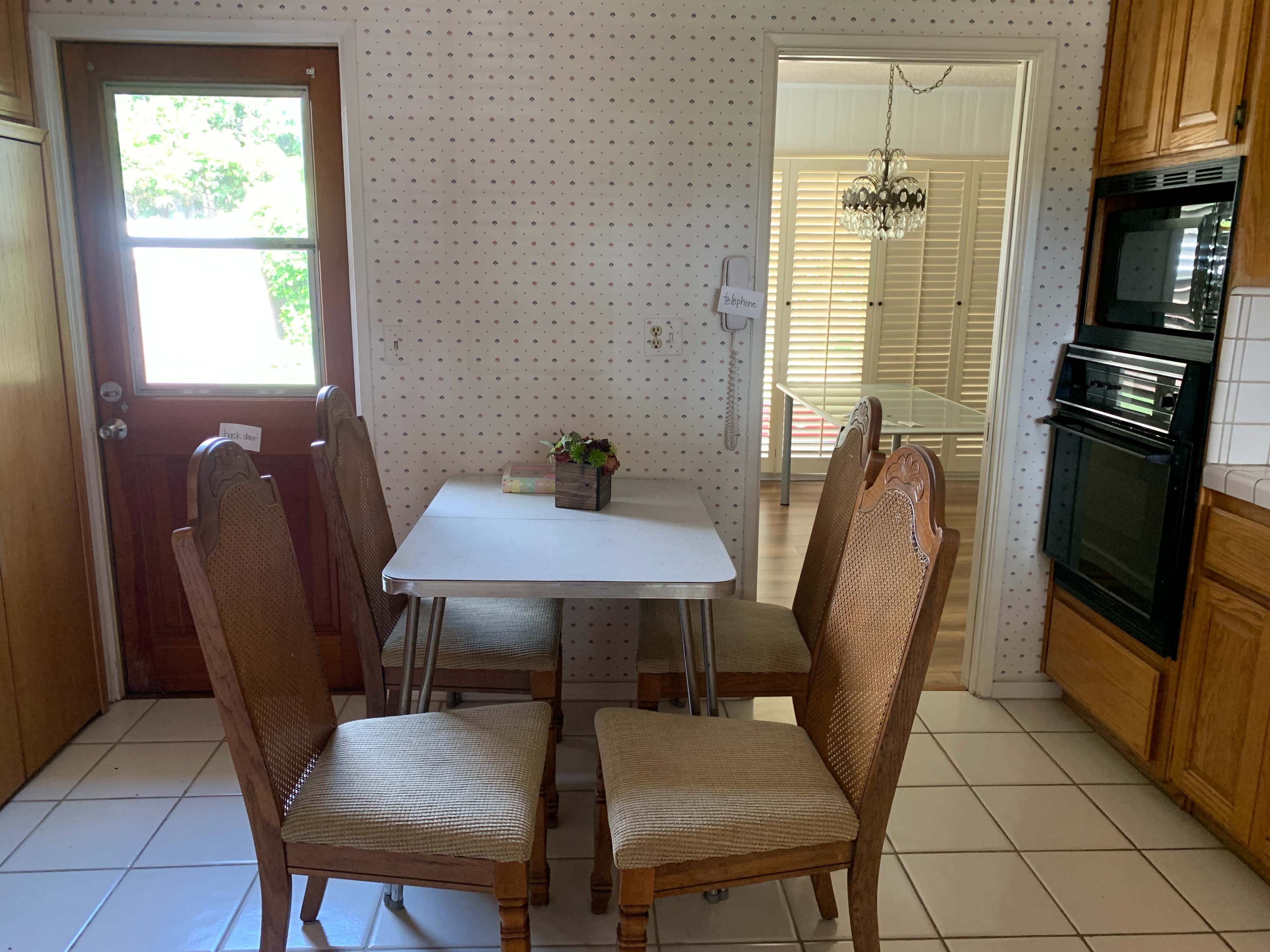 A small kitchen features a white table surrounded by four wooden chairs, with an adjacent doorway leading to another room.