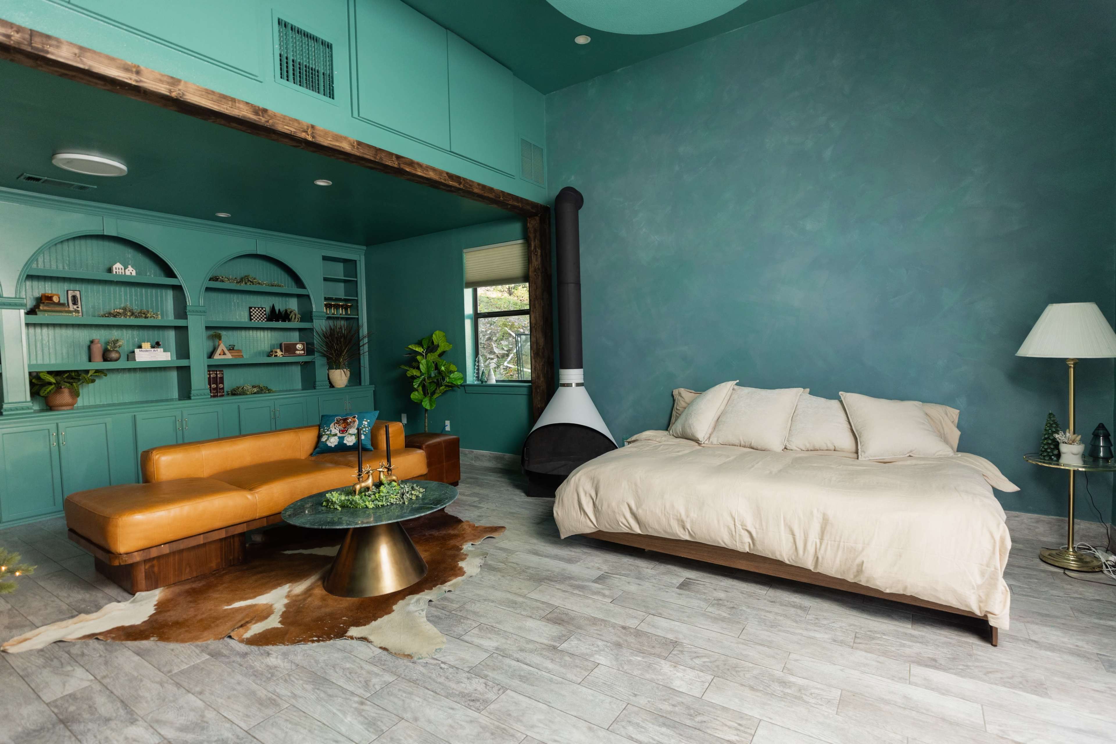 A room with teal walls, a brown leather couch, a beige bed, a circular green coffee table, and a modern fireplace.
