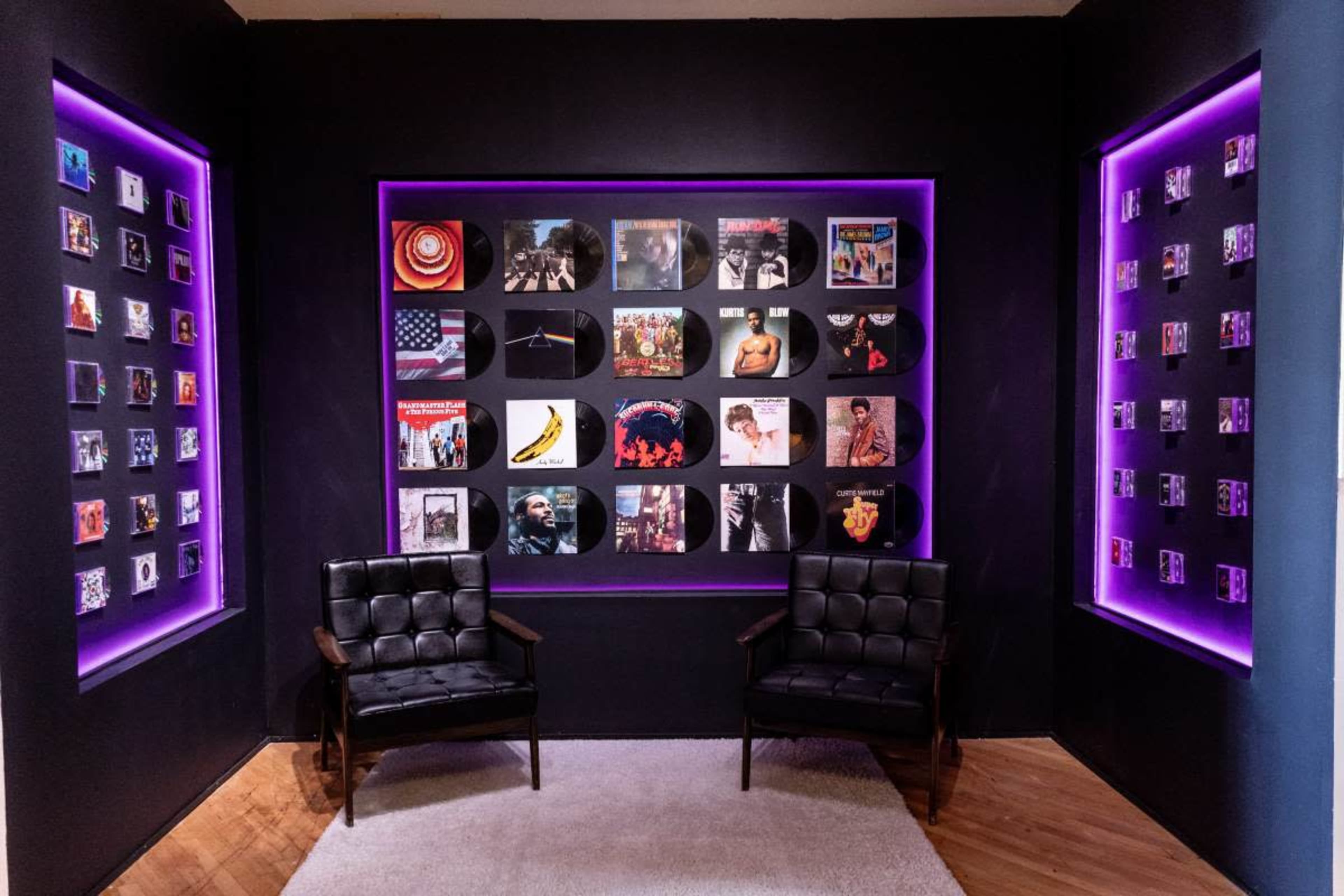 The image features a stylish room with two black chairs facing a wall displaying various vinyl records framed with purple lighting.