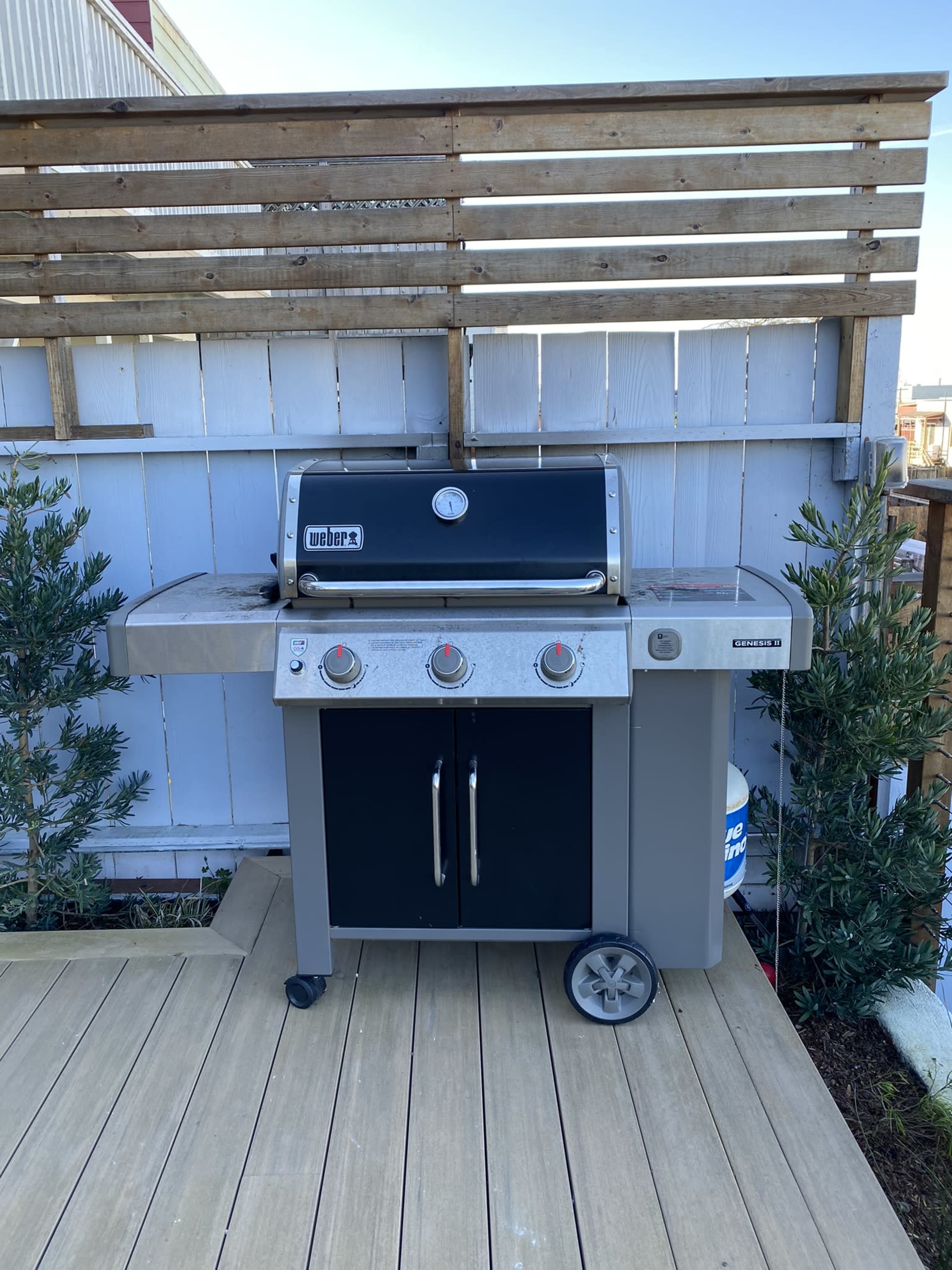 A gas grill situated on a wooden patio, positioned against a white wooden fence with slats.