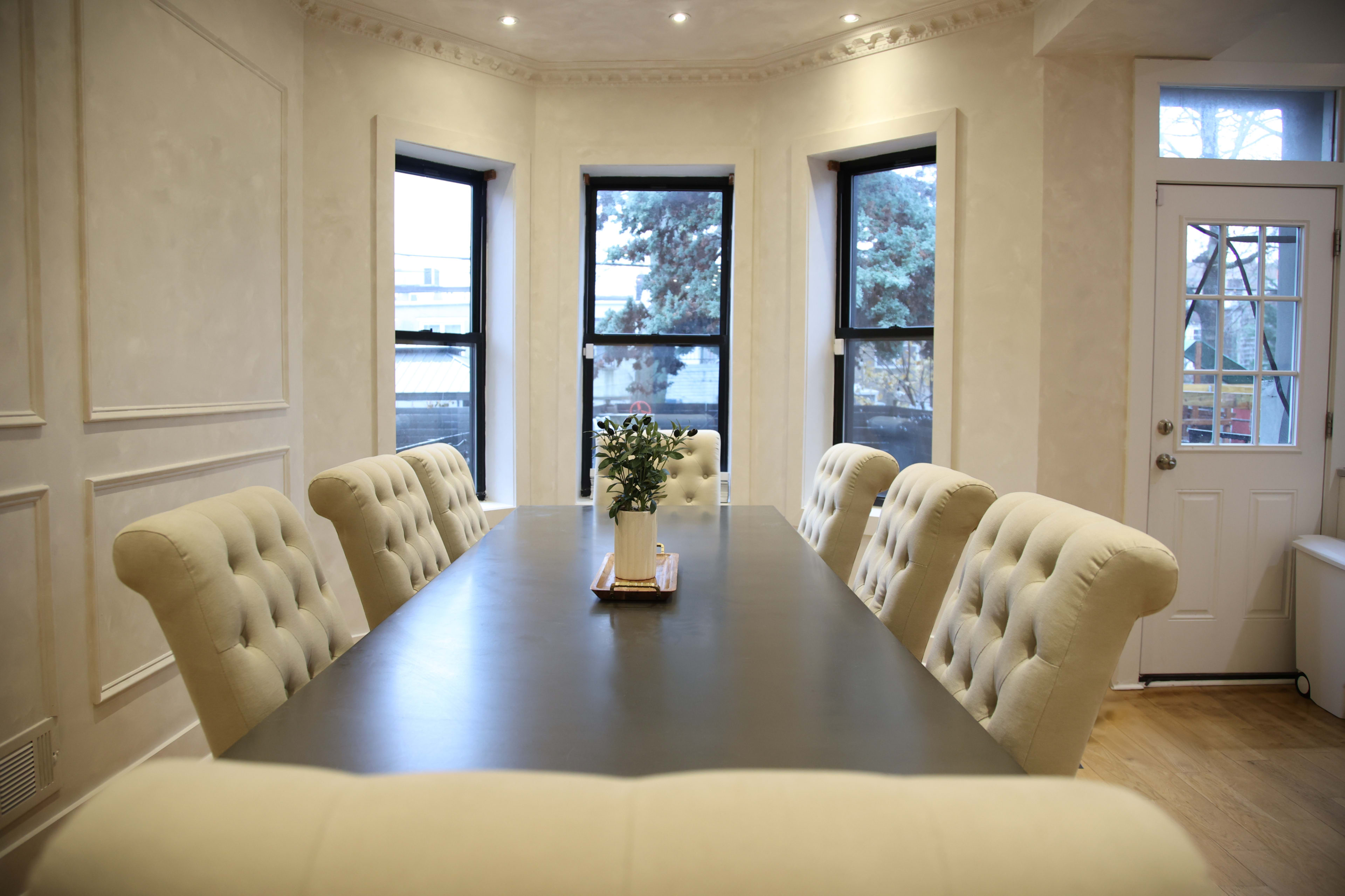 A long rectangular table with upholstered chairs is set in a well-lit dining room featuring large windows and a door.