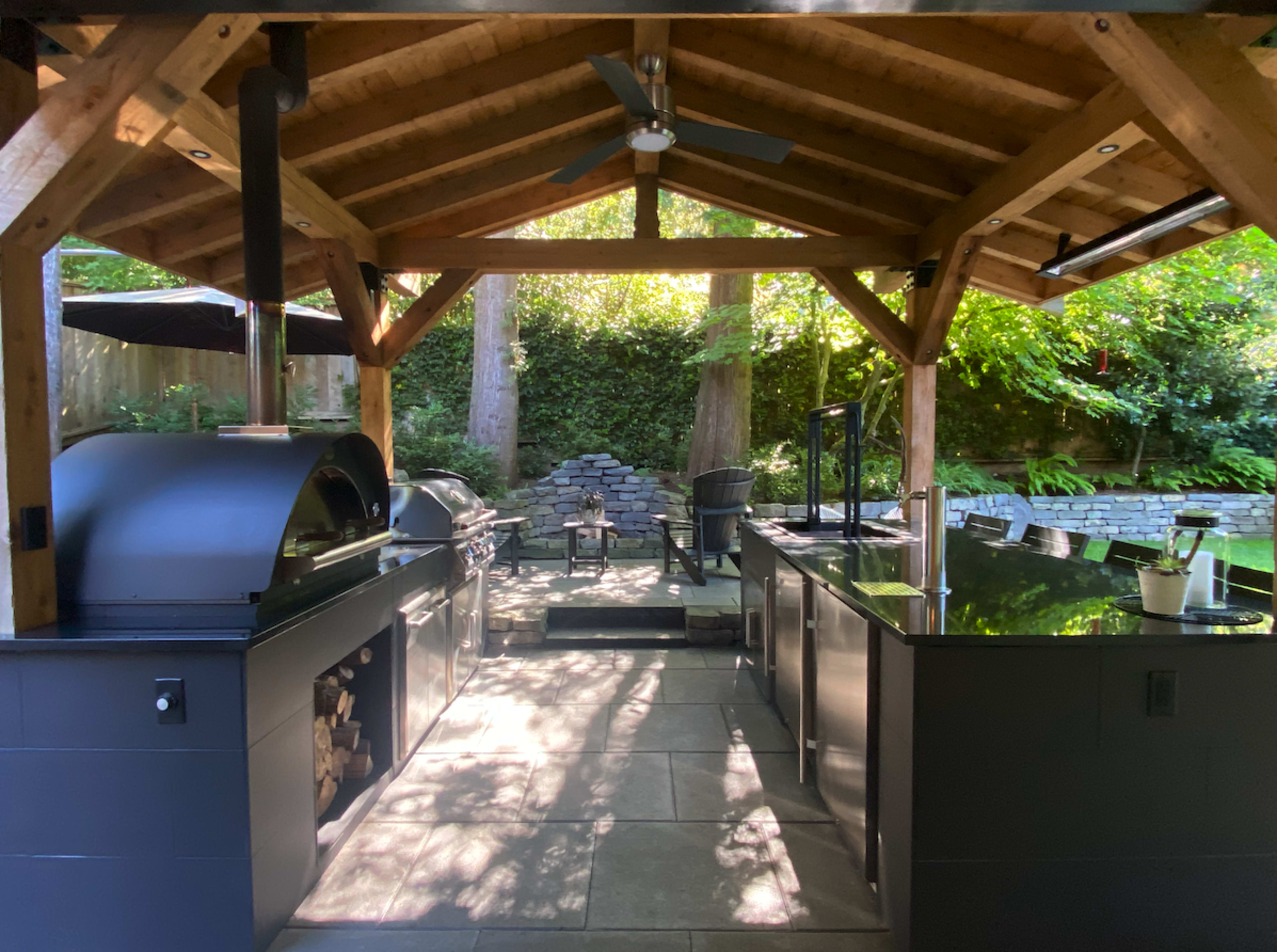 The image shows an outdoor kitchen under a wooden structure, equipped with a grill, pizza oven, and counter space, surrounded by greenery.