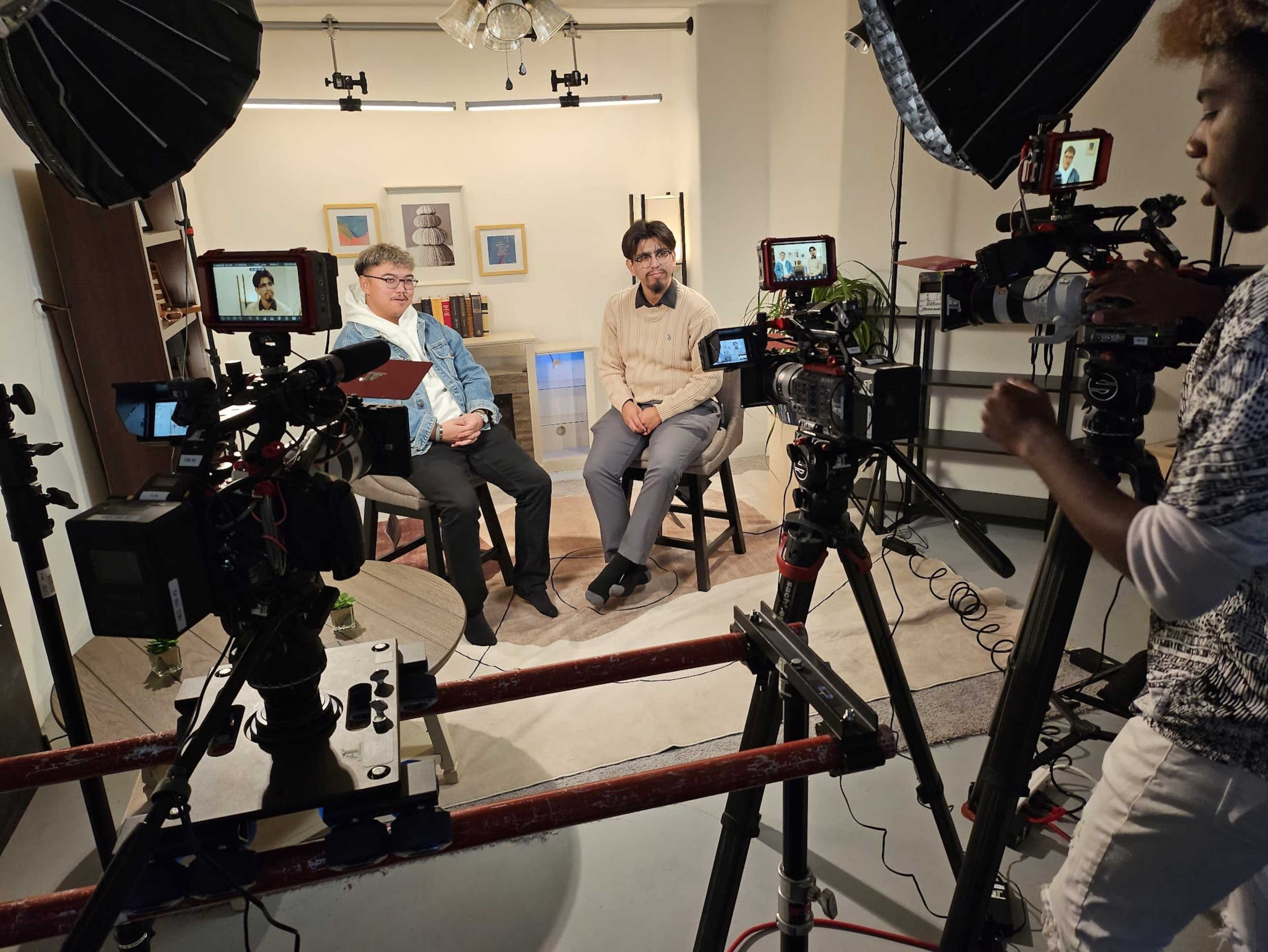 Two men sit in chairs for an interview, surrounded by multiple cameras and lighting equipment in a studio setting.
