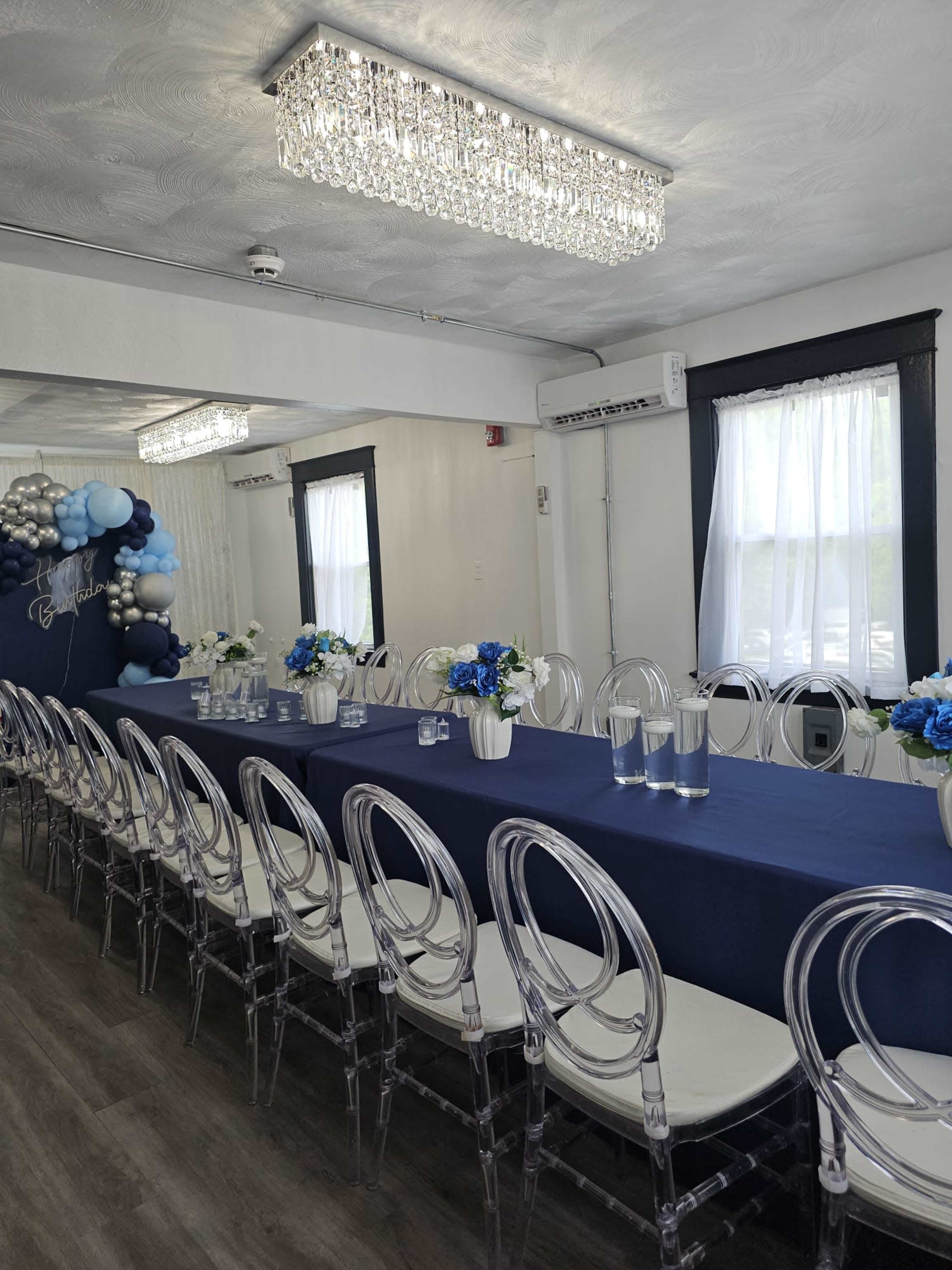 The image shows a banquet room with a long table covered in a blue tablecloth, surrounded by clear acrylic chairs, and decorated with white and blue floral centerpieces.