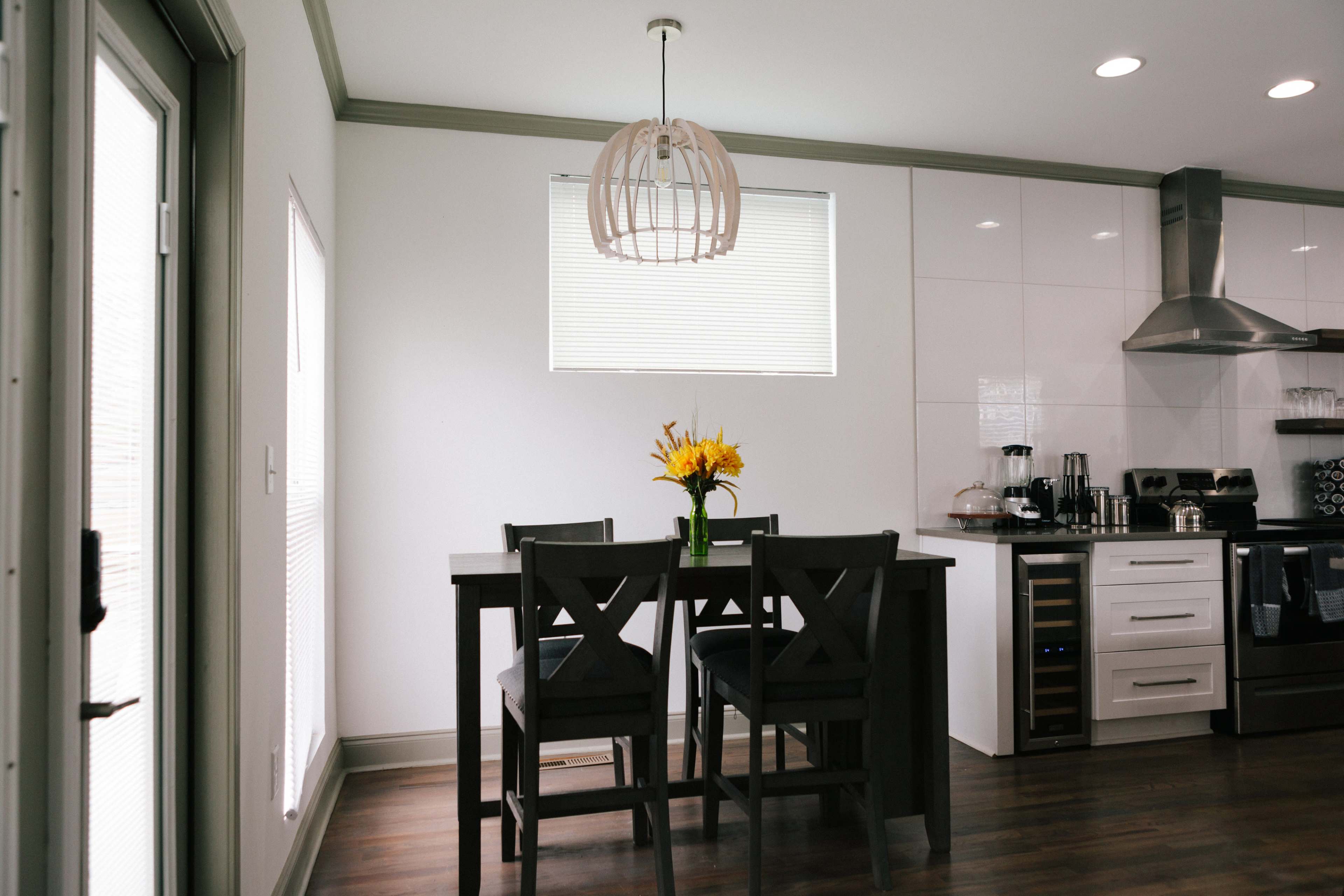 A modern kitchen with a dining table, four chairs, and a vase of yellow flowers, illuminated by a pendant light.