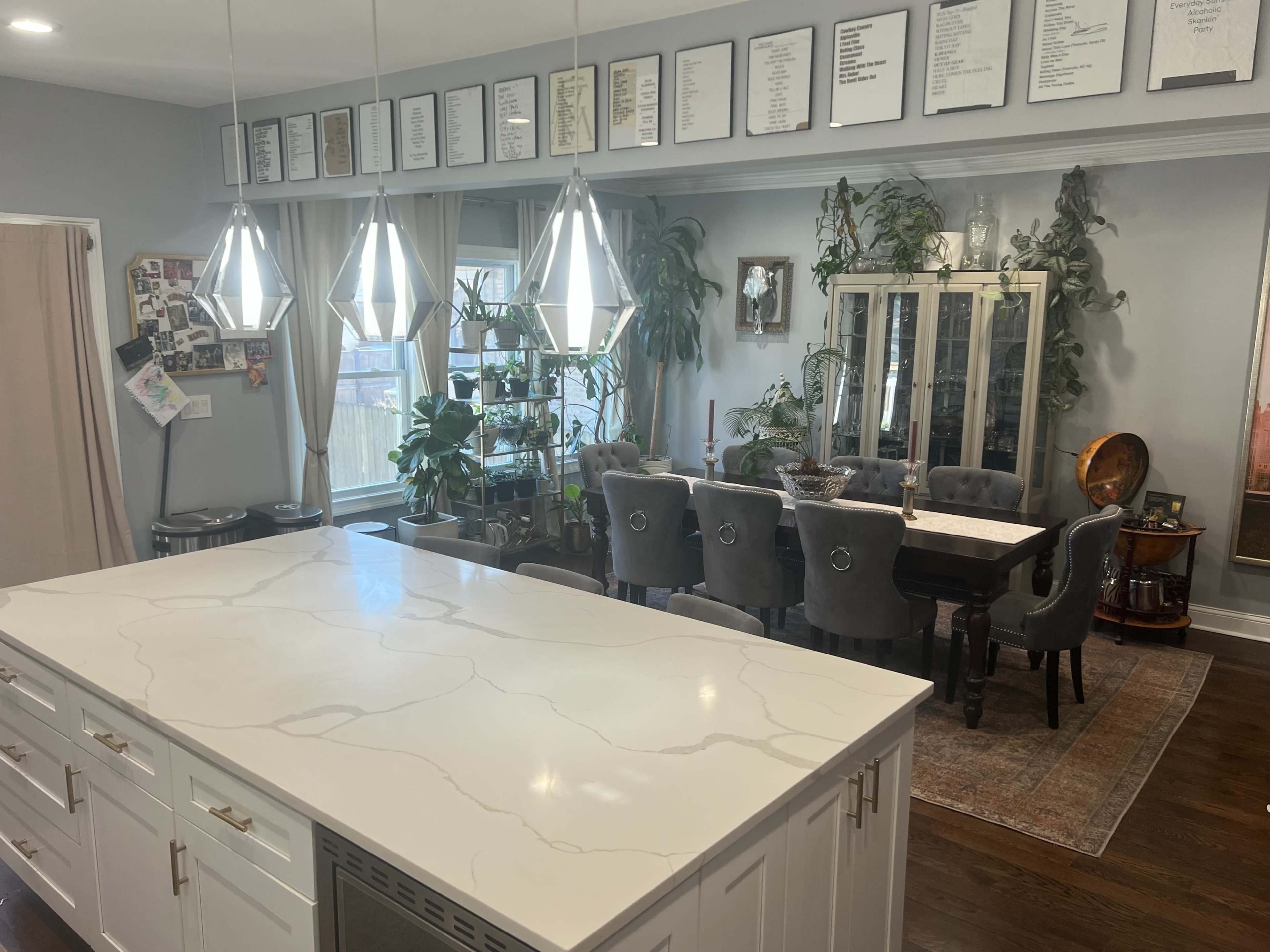 A modern kitchen with a large island and pendant lighting opens up to a dining area featuring a glass cabinet and a long dining table surrounded by upholstered chairs.