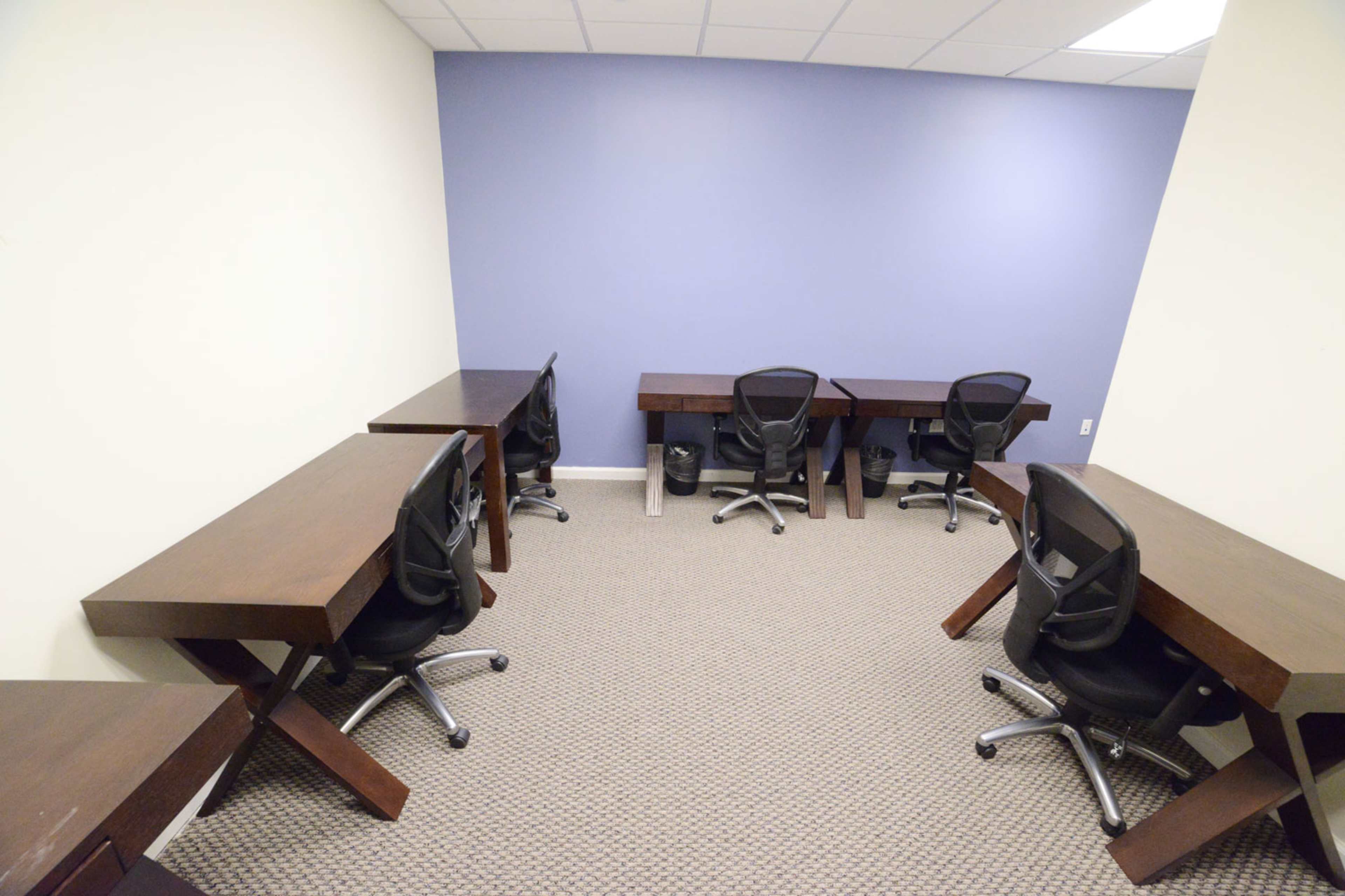 The image shows a small office space with six wooden desks and black mesh chairs arranged along the walls of a room featuring a light-colored wall and a purple accent wall.