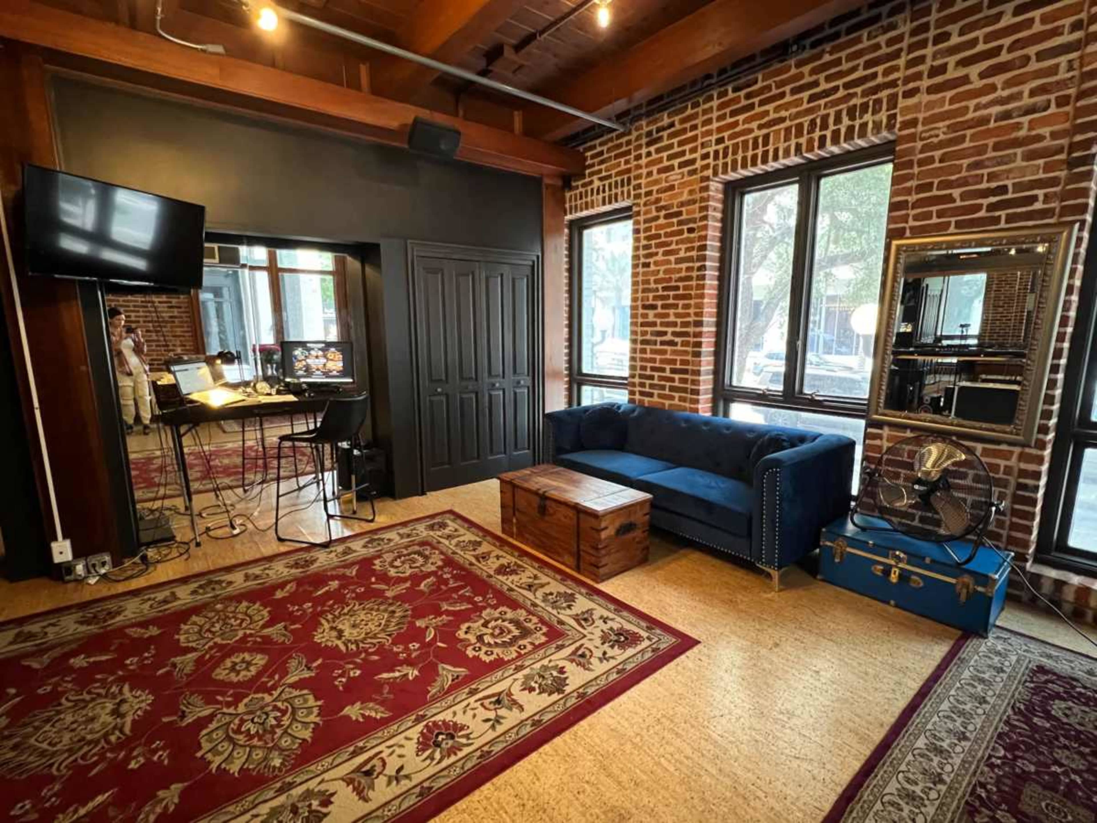 The image shows a modern living space with exposed brick walls, a blue couch, a wooden coffee table, and a large TV mounted on the wall, all arranged on a patterned area rug.