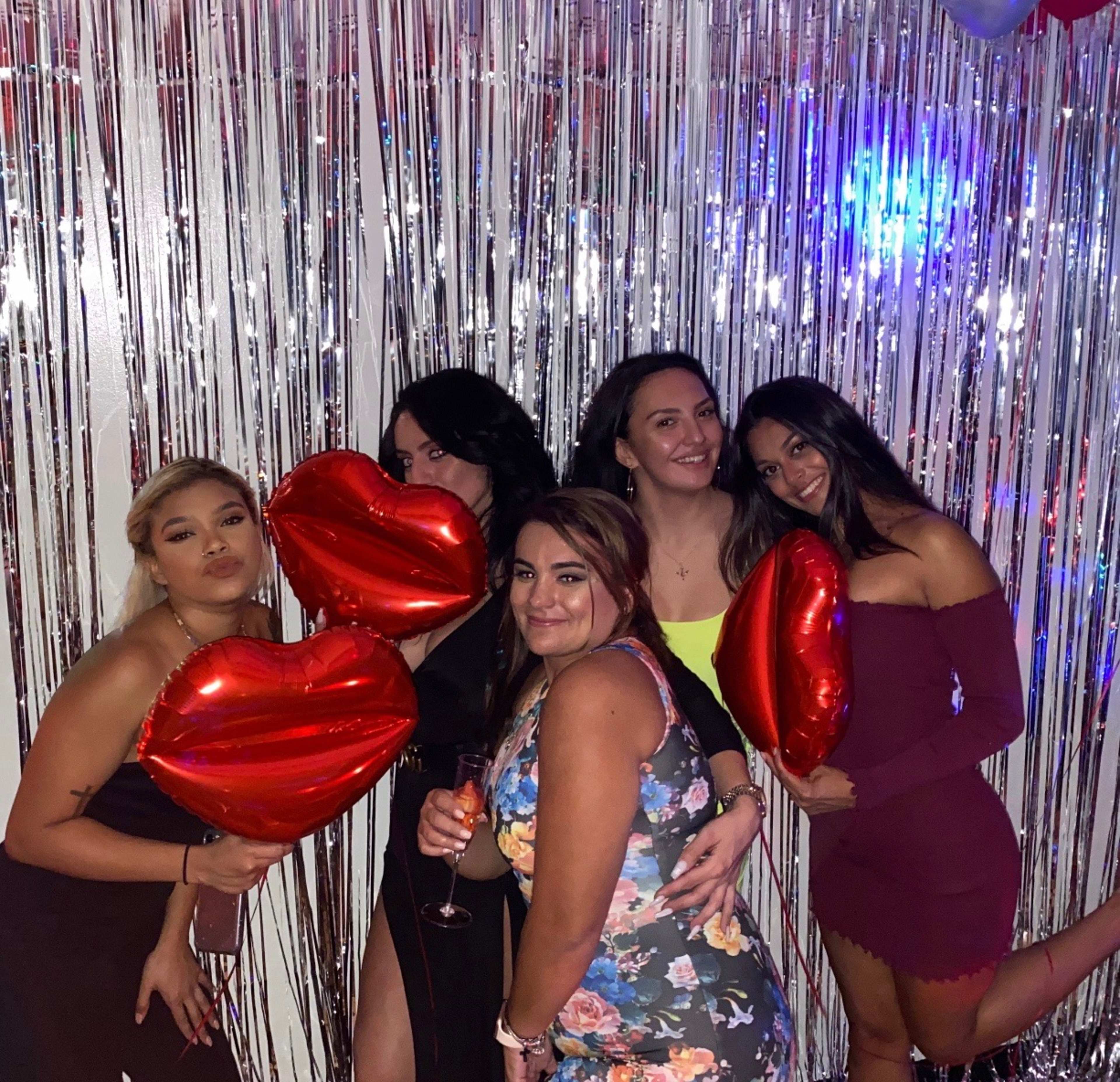 A group of five women poses with large red heart-shaped balloons in front of a shiny silver backdrop.