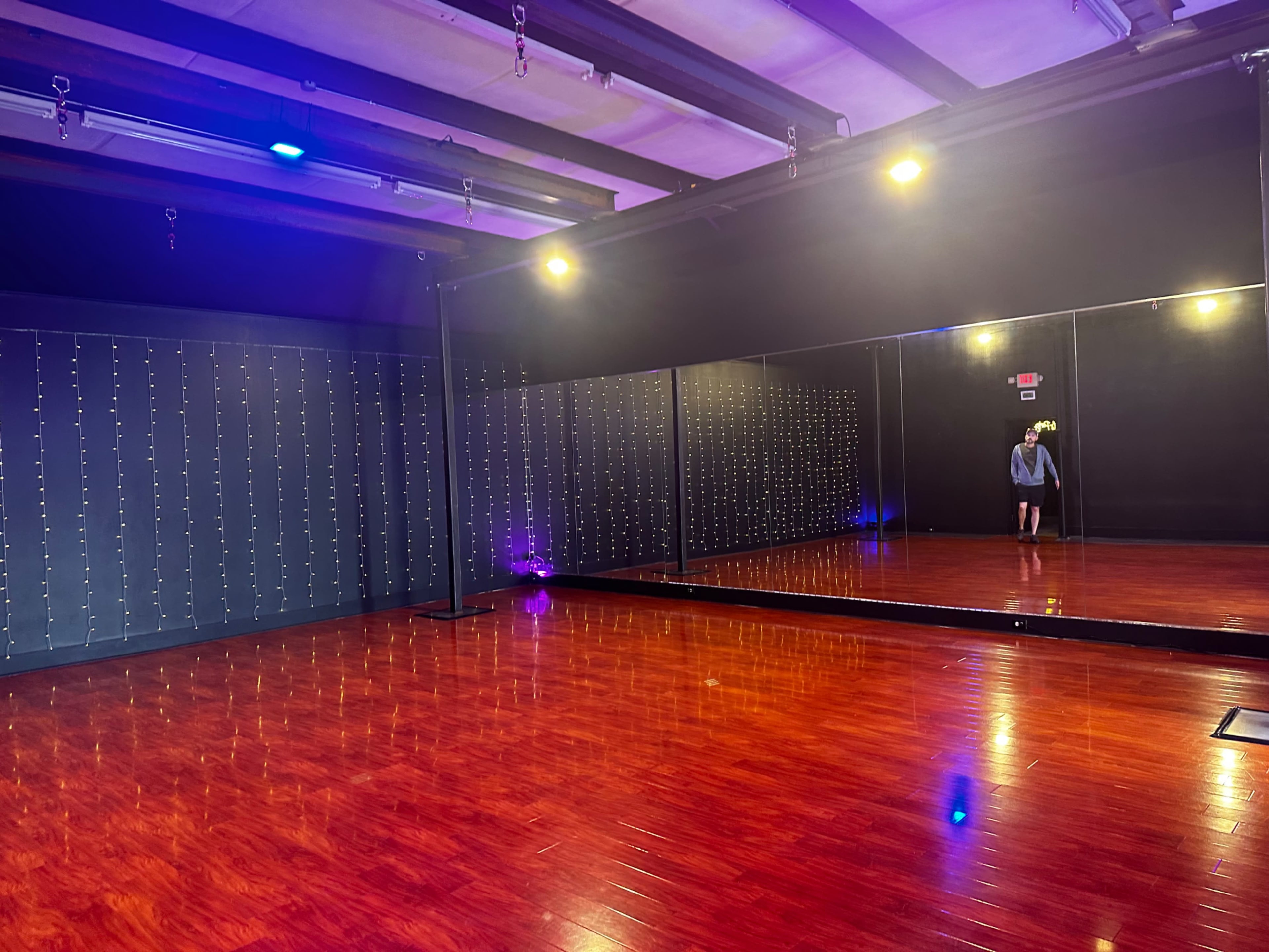 A dance studio with a polished wooden floor, black walls, string lights, and a large mirror reflecting a person standing in the doorway.
