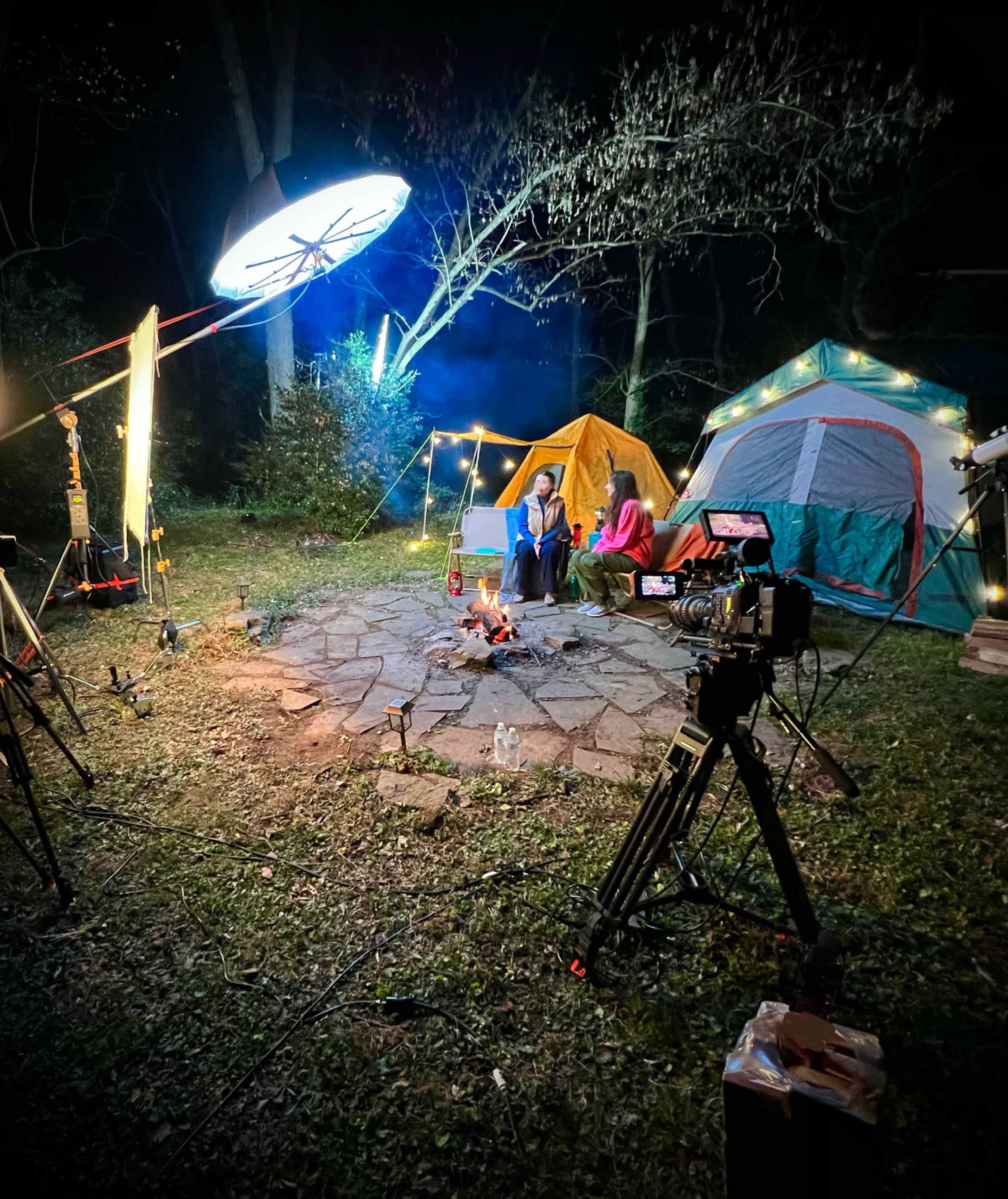 The scene shows two people sitting by a campfire surrounded by tents and film equipment in a nighttime outdoor setting.