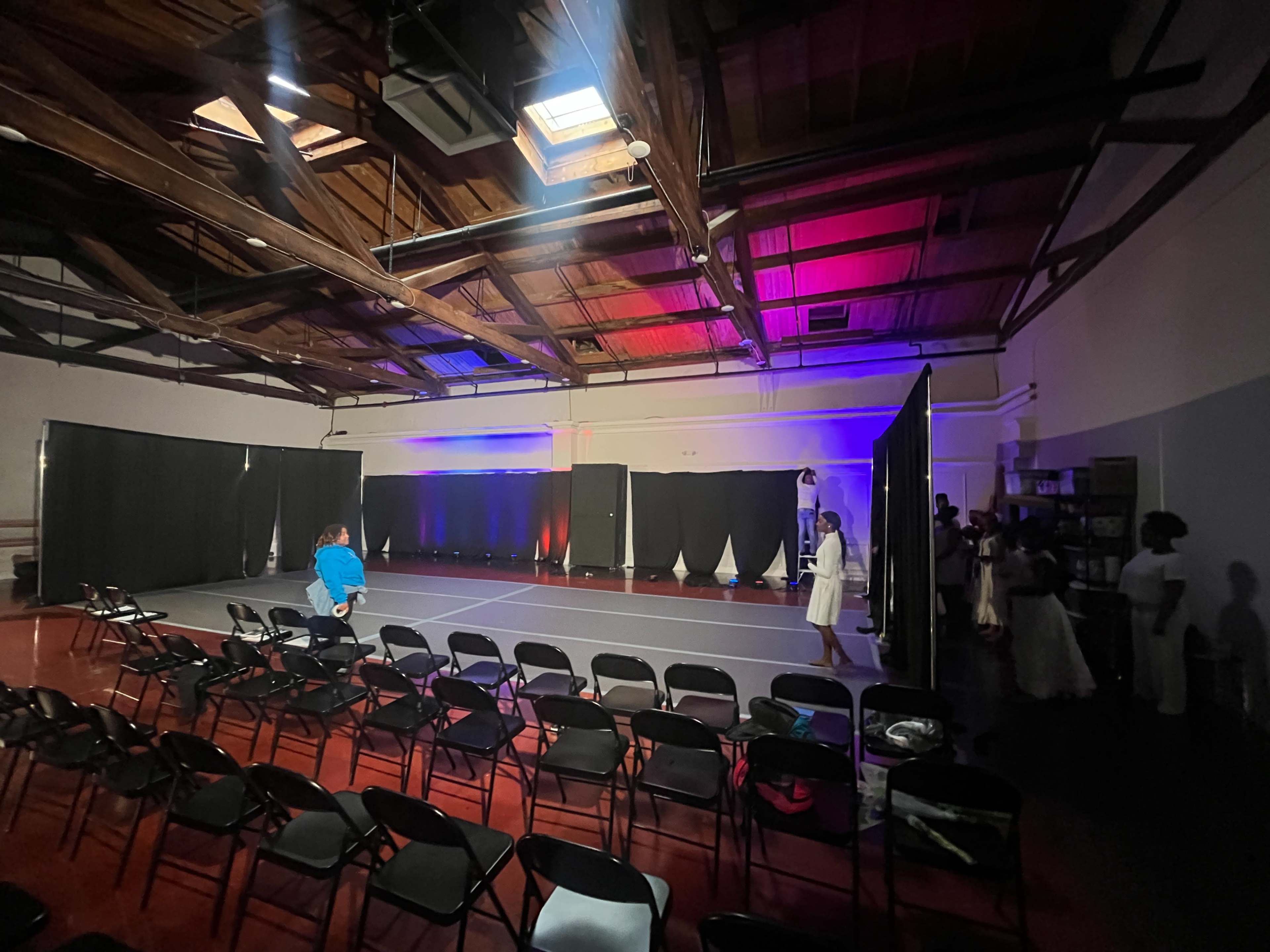 A spacious performance venue is set up with rows of black chairs facing a stage that is framed by black curtains and illuminated with colored lights.