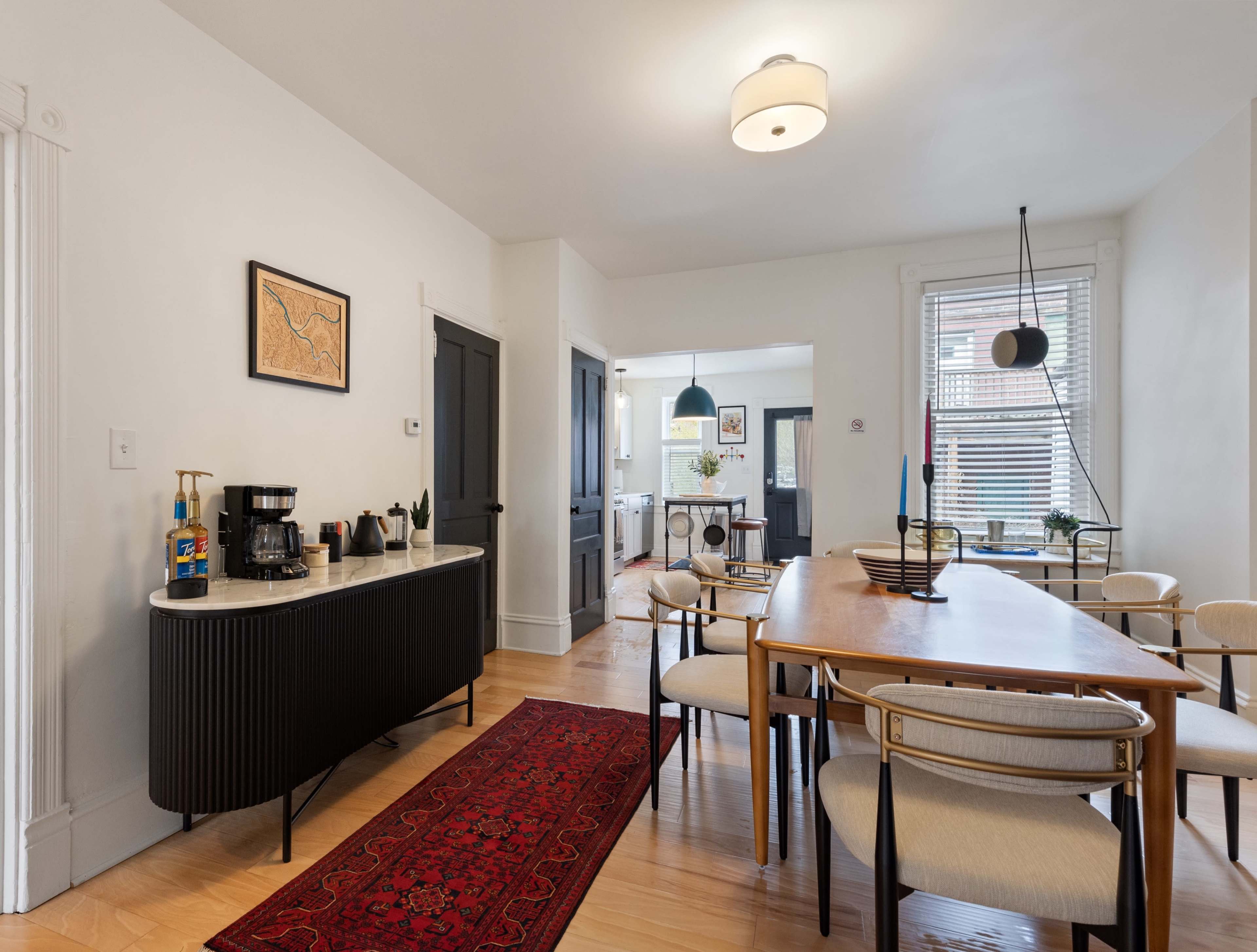 A modern dining area features a wooden table surrounded by chairs, with a sideboard displaying a coffee maker and plants, against a backdrop of light-colored walls and hardwood floors.