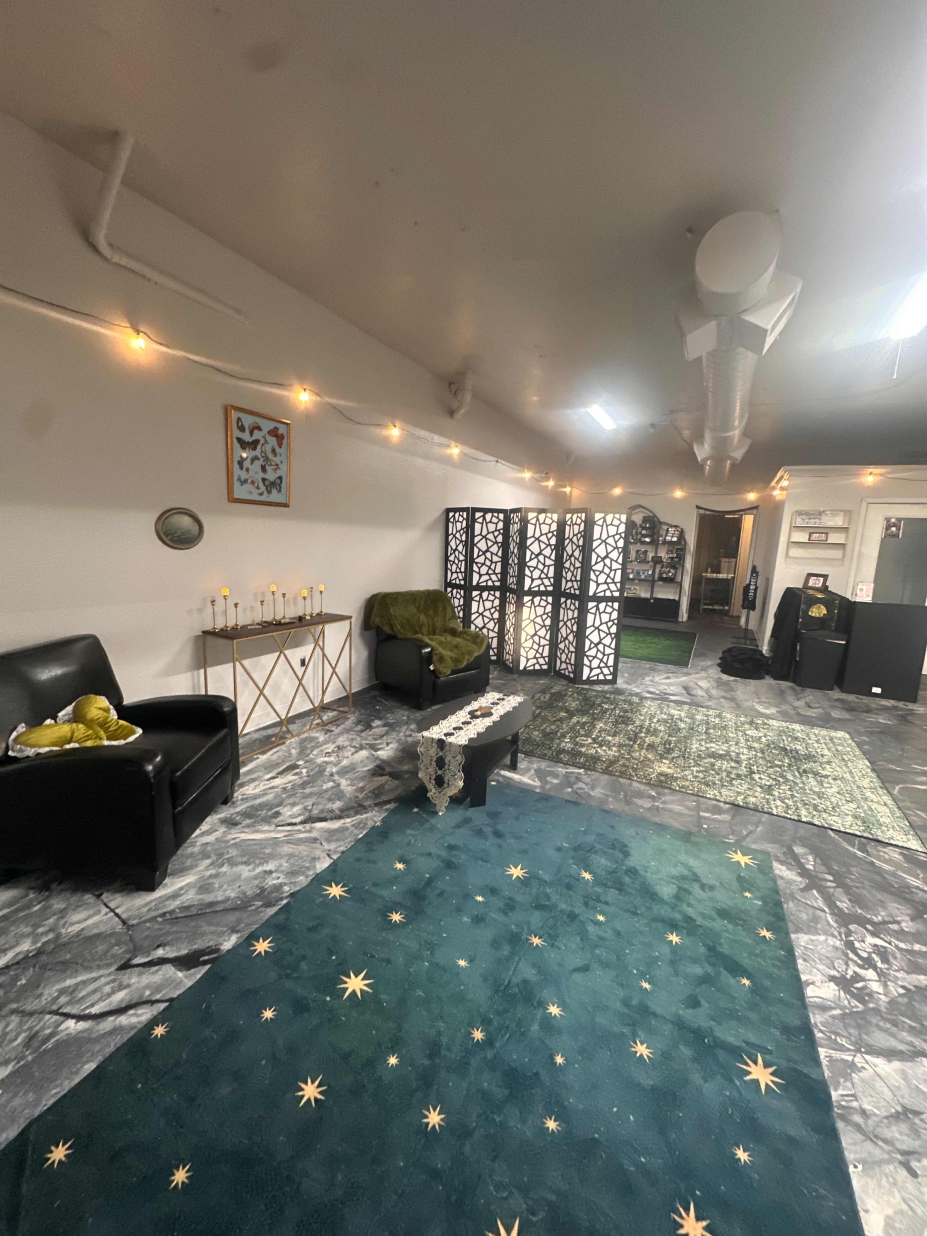 The image depicts a spacious room with dark flooring, featuring two black chairs, a decorative screen, various rugs, candles on a table, and soft lighting.