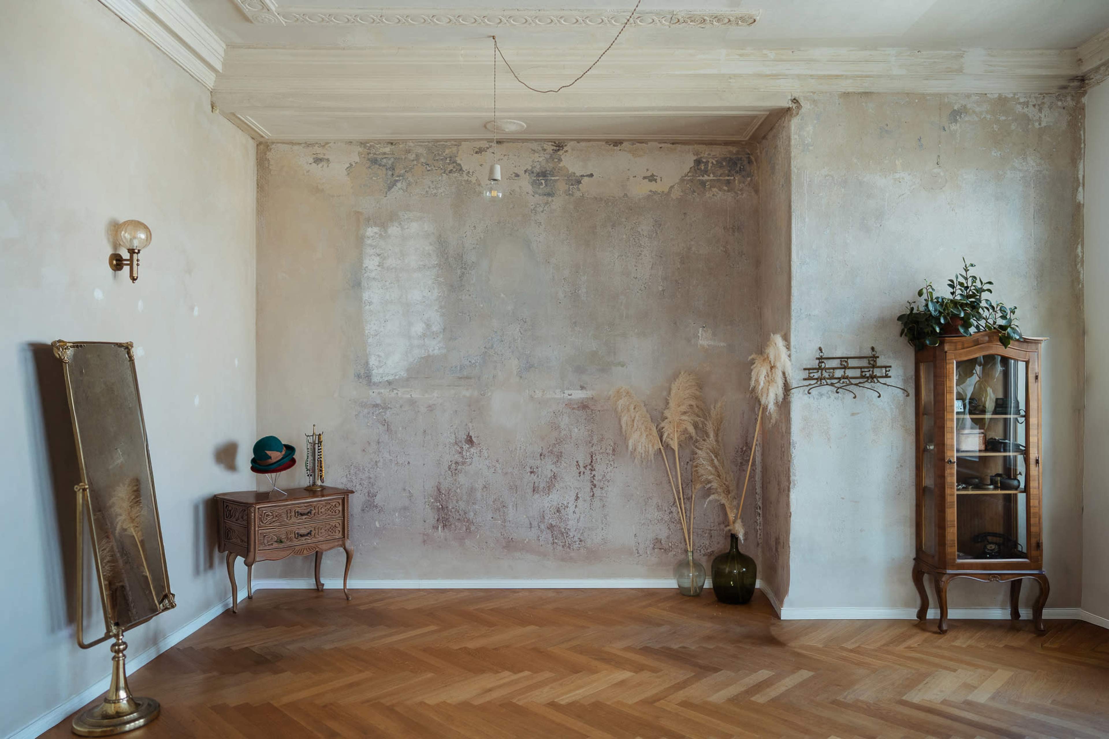 The image shows a minimalist room with a wooden floor, featuring a vintage dresser, a wall-mounted cabinet, decorative plants, a large mirror, and peeling paint on the walls.
