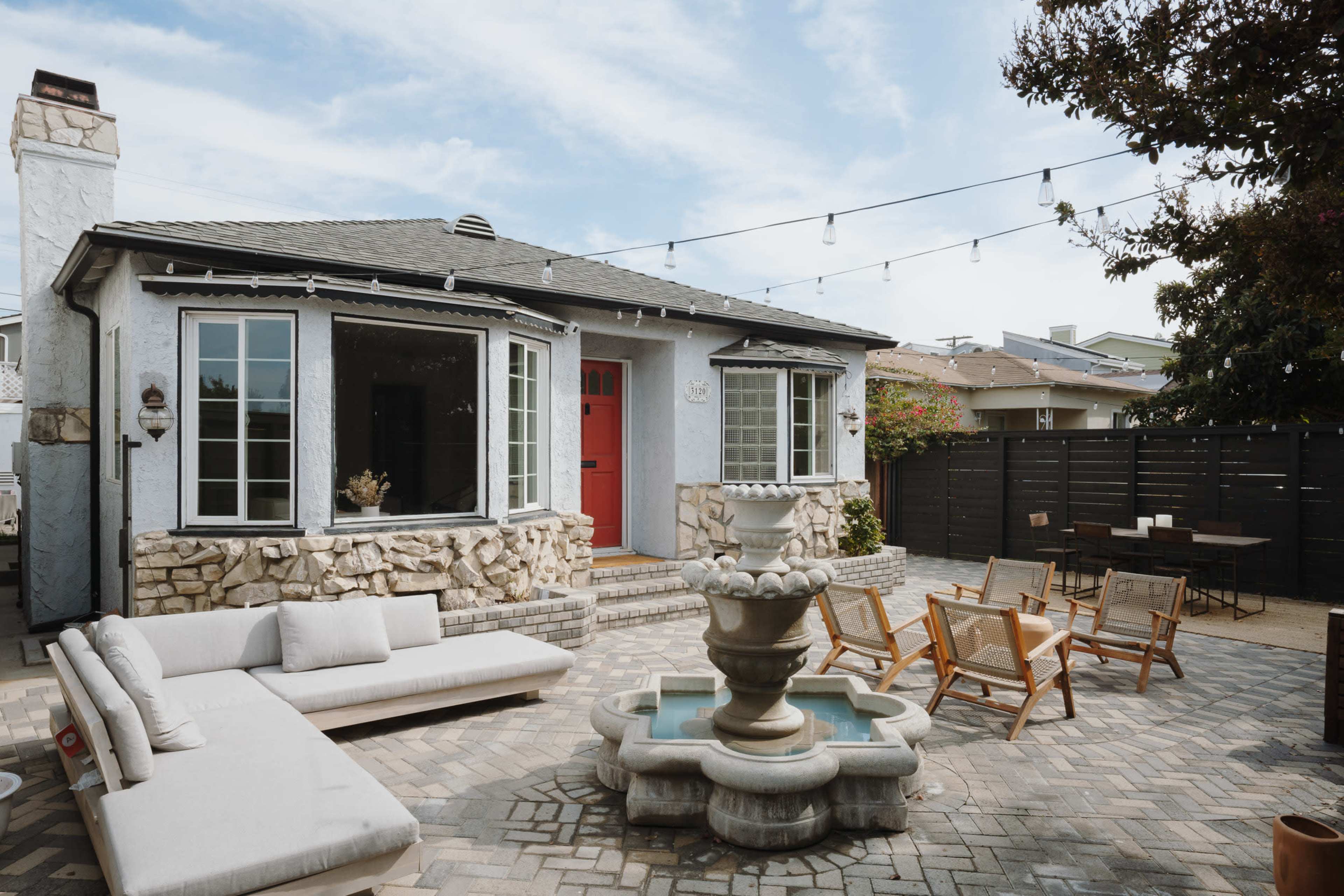 A cozy backyard features a stone fountain surrounded by seating areas and a house with a red door.
