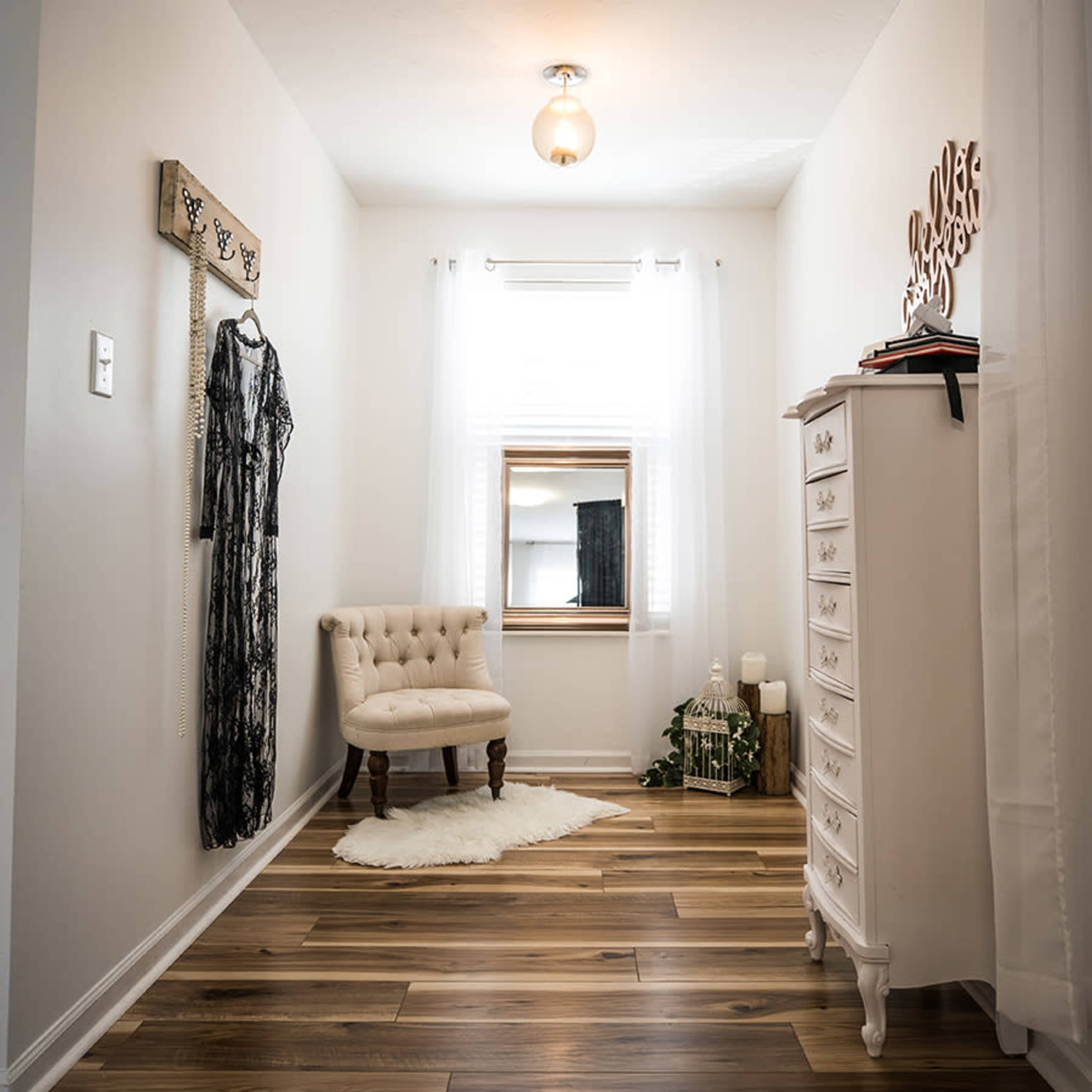 A bright room with light-colored walls, wooden flooring, a tufted chair, a decorative mirror, and a white dresser positioned against the wall.