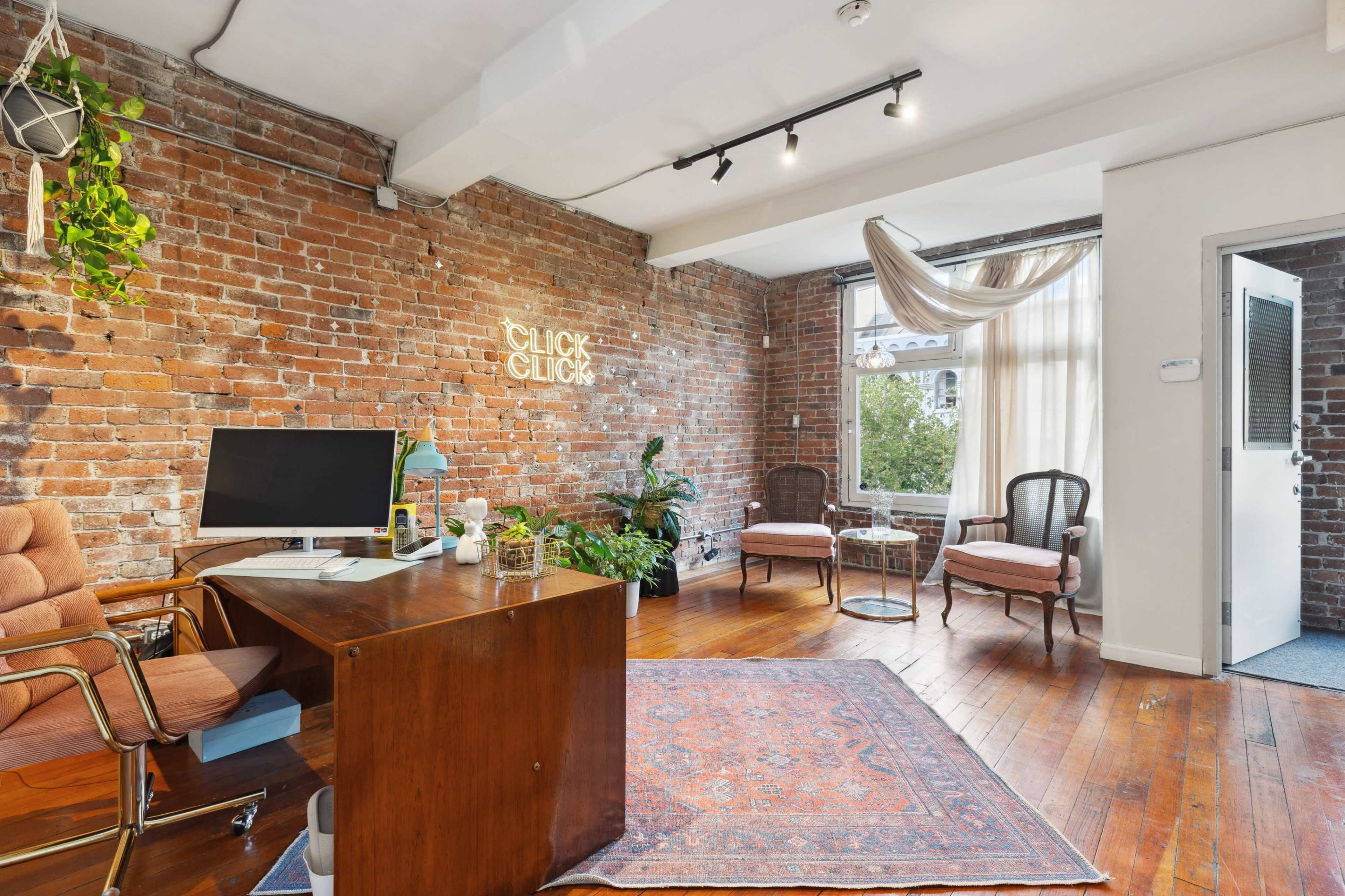 The scene features a stylish office space with brick walls, wooden flooring, a desk with a computer, and two chairs near a window adorned with drapes.