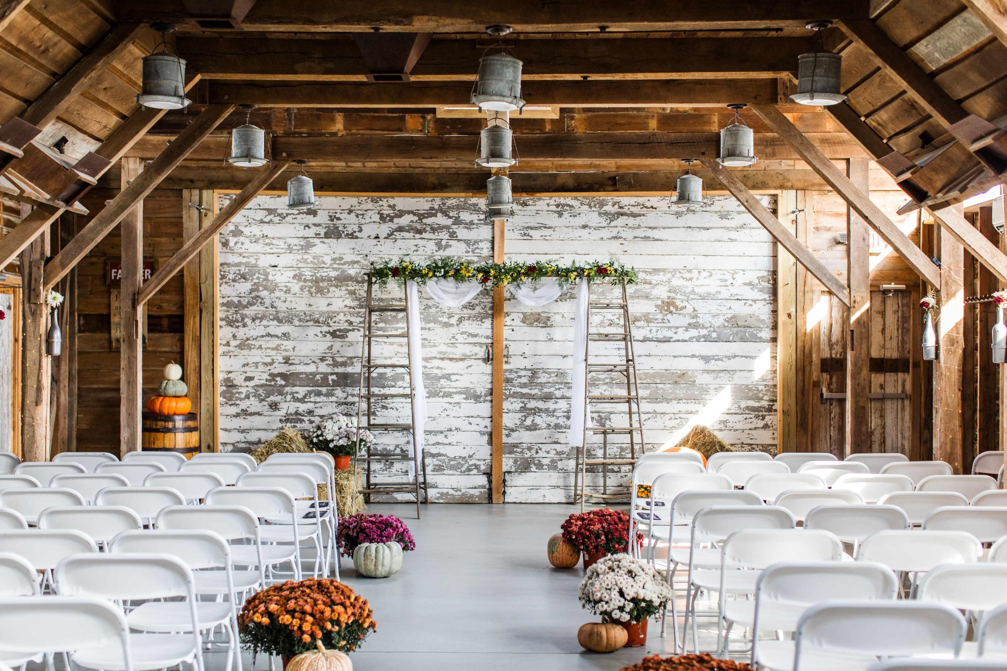 An indoor venue features rows of white chairs facing a decorative arch made of ladders adorned with flowers, surrounded by pumpkins and potted flowers.
