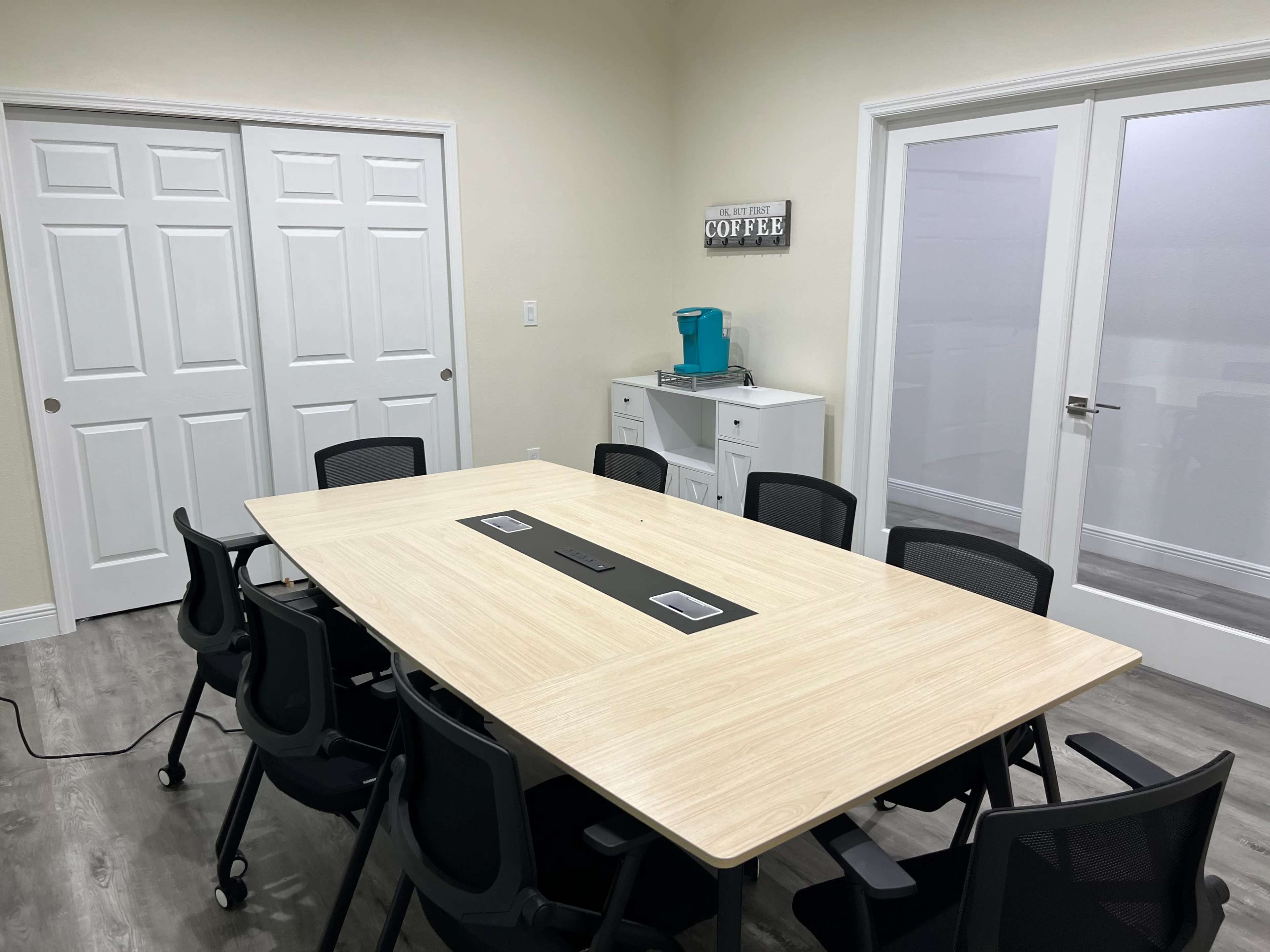 The image shows a conference room with a large rectangular table surrounded by black rolling chairs, a coffee station on one side, and double doors leading to another room.