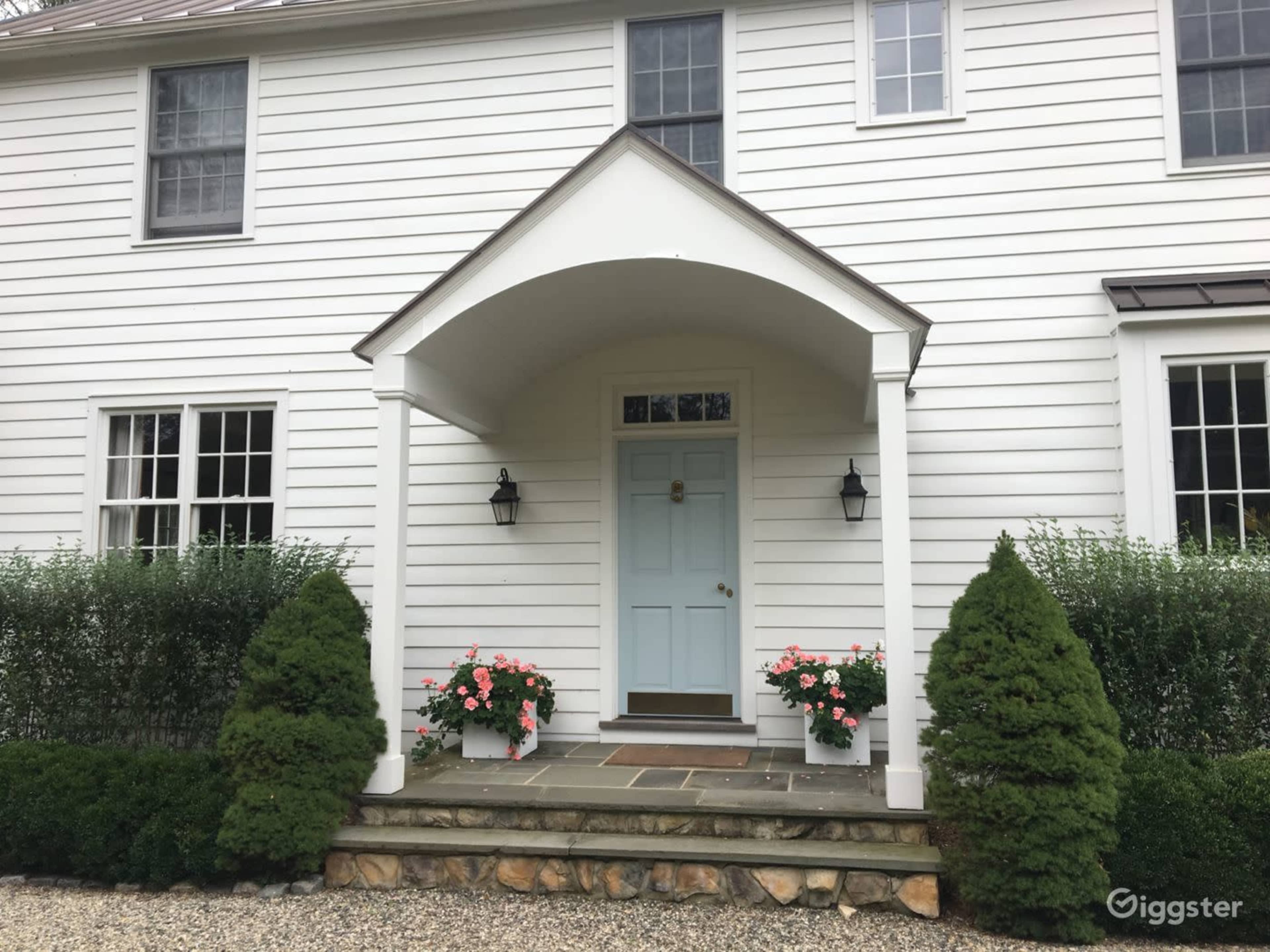 The image shows the entrance of a two-story white house with a covered porch, blue front door, and flower pots on either side of the steps.