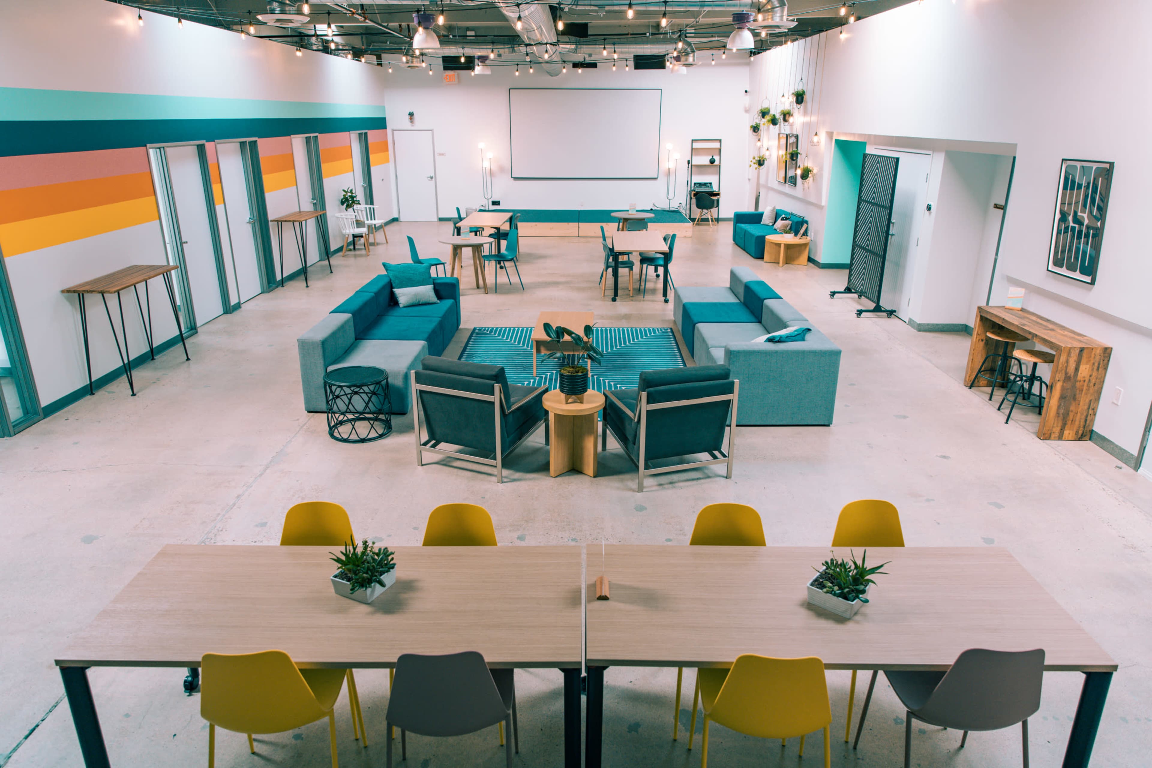 A spacious, modern interior with various seating arrangements, including tables and chairs, surrounded by colorful wall stripes and decorative plants.