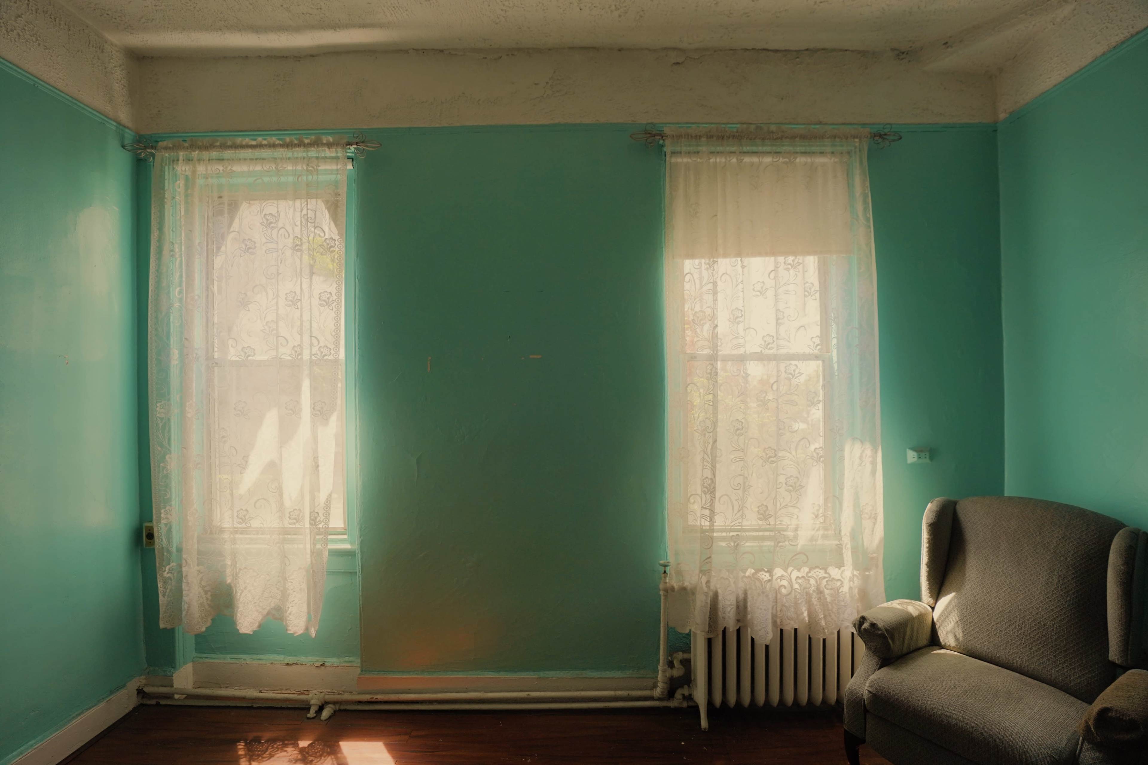 A room with turquoise walls, two windows dressed with lace curtains, and a gray armchair in the corner.