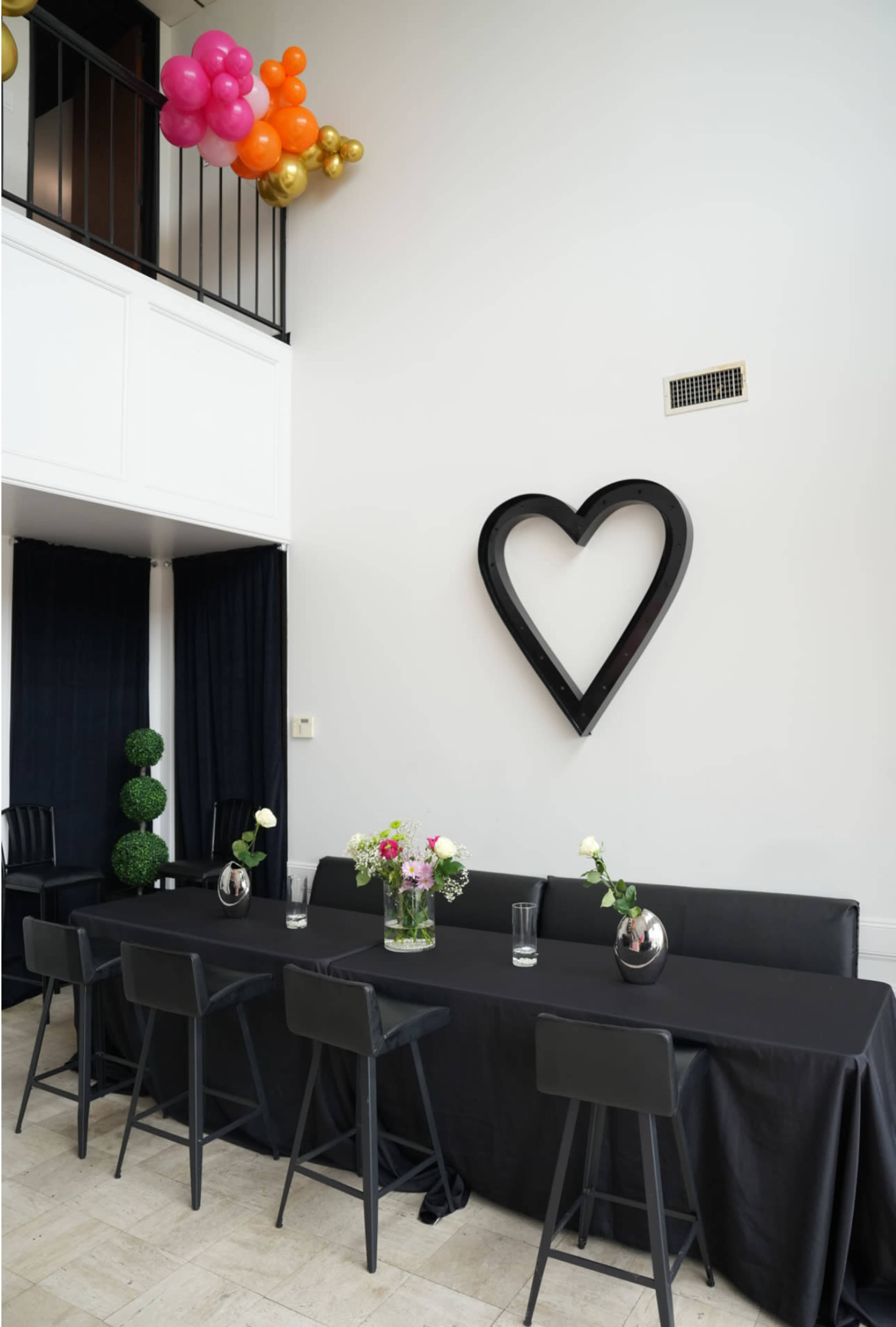 The image shows a decorated event space with a long black table set for guests, surrounded by black chairs, and adorned with flowers and a large heart-shaped wall decoration.