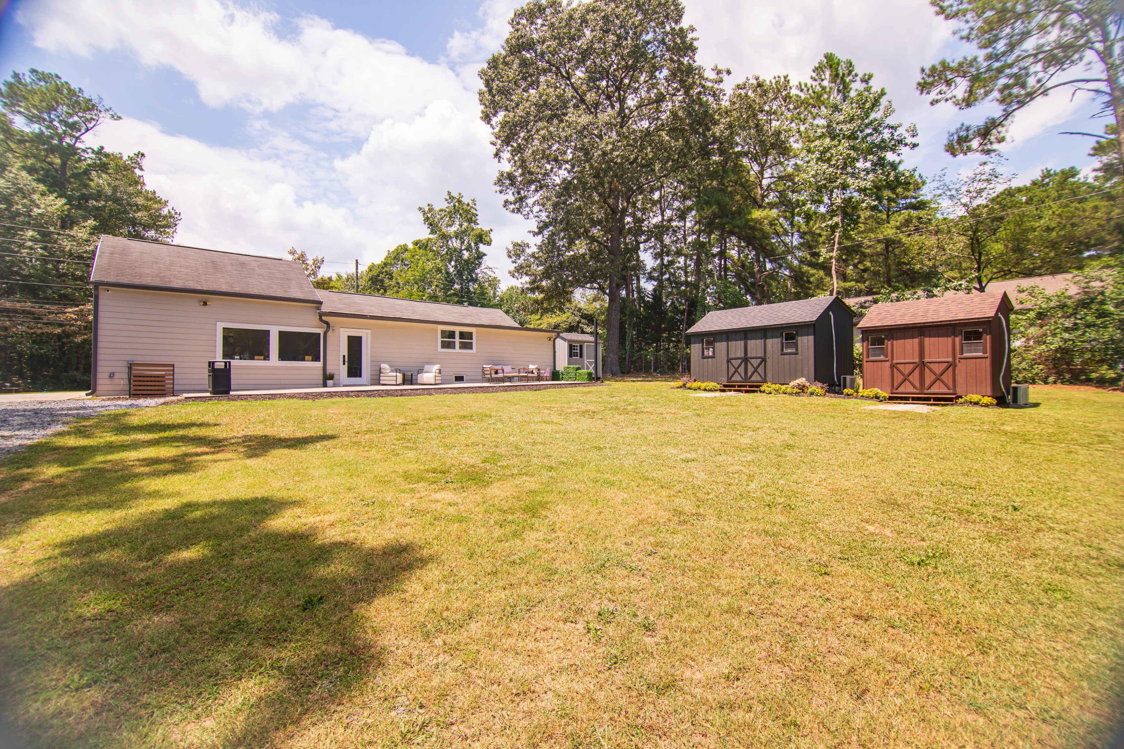 The image shows a well-maintained yard with a single-story house on the left and two storage sheds on the right, surrounded by trees and open grass.