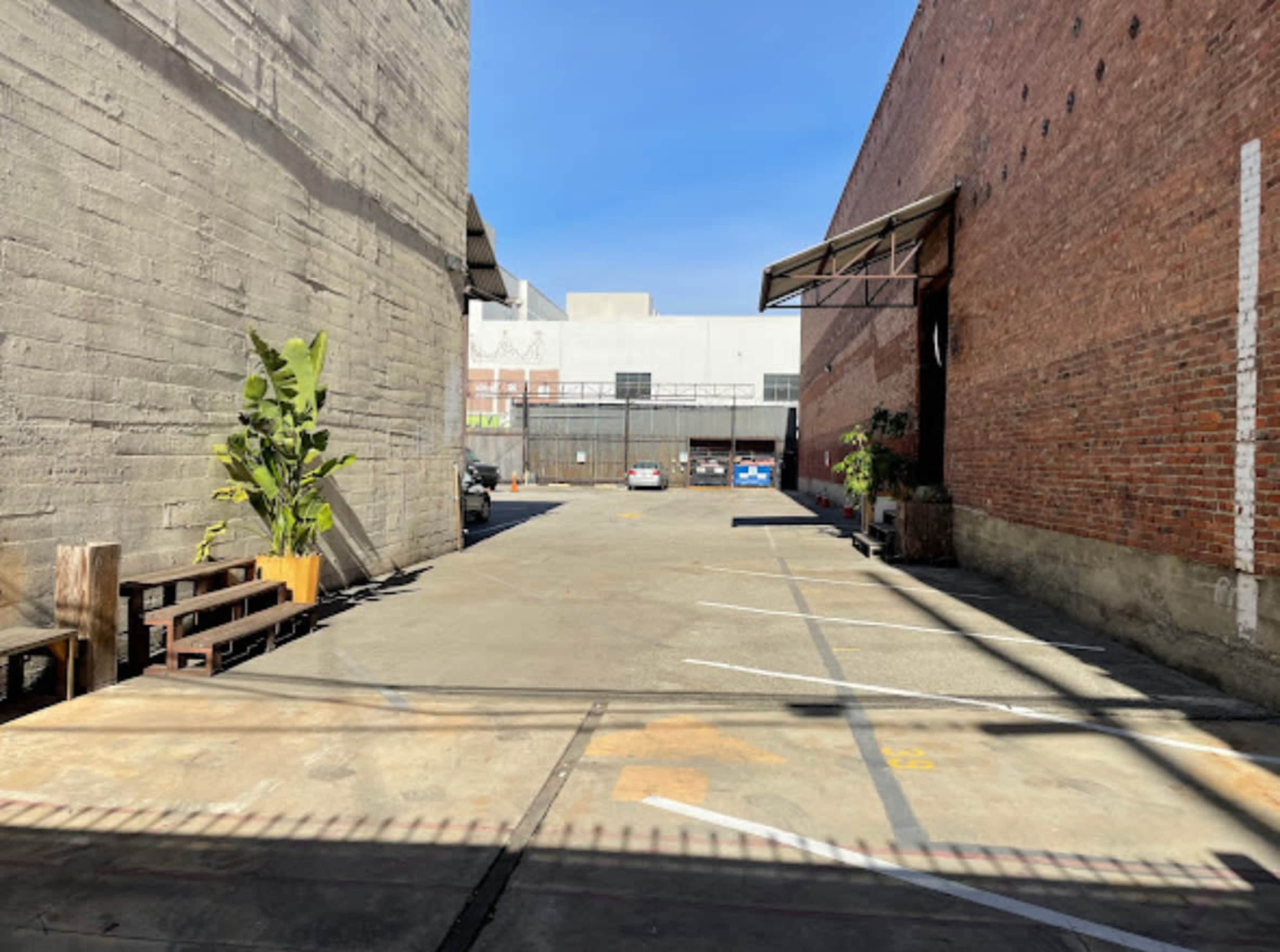 The image shows a vacant parking lot situated between two industrial buildings, with potted plants and a few parked cars visible.