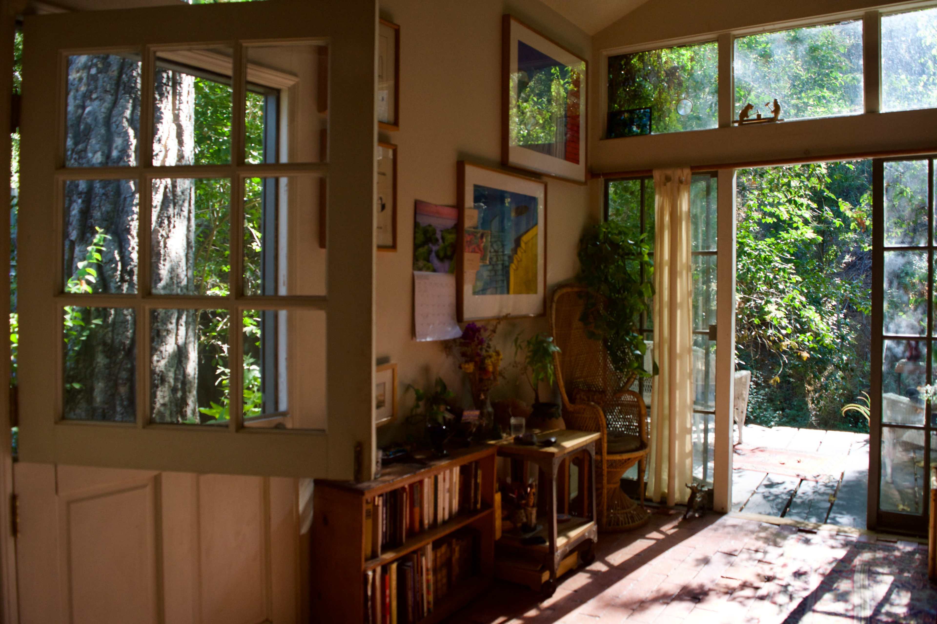 A sunlit room opens to a garden through large glass doors, featuring a wooden table, bookshelves, and framed artwork on the walls.