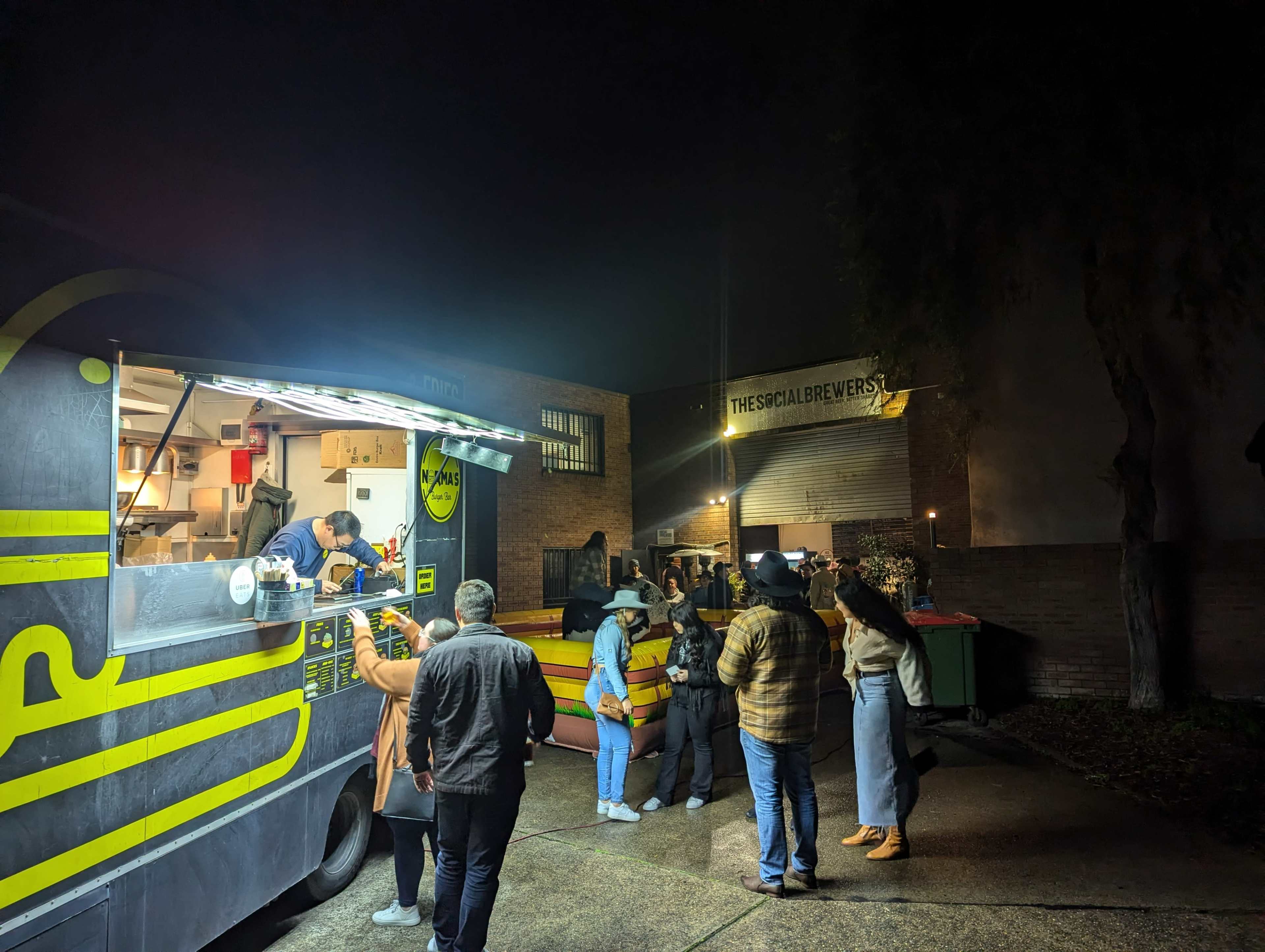 A food truck is serving a line of customers outside a brewery at night.
