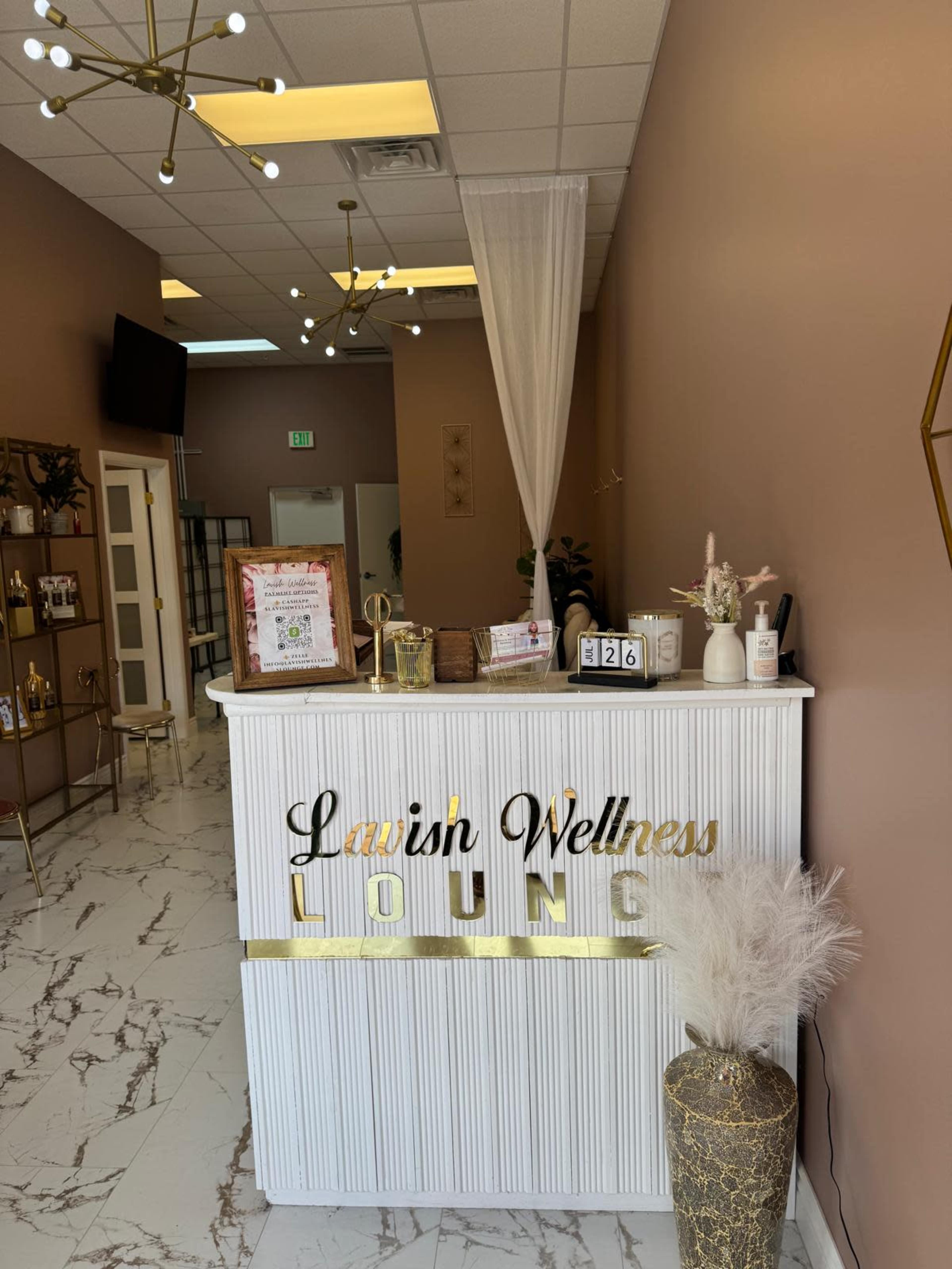 The image shows a reception area of a wellness lounge with a white desk, decorative elements, and a neutral color palette.