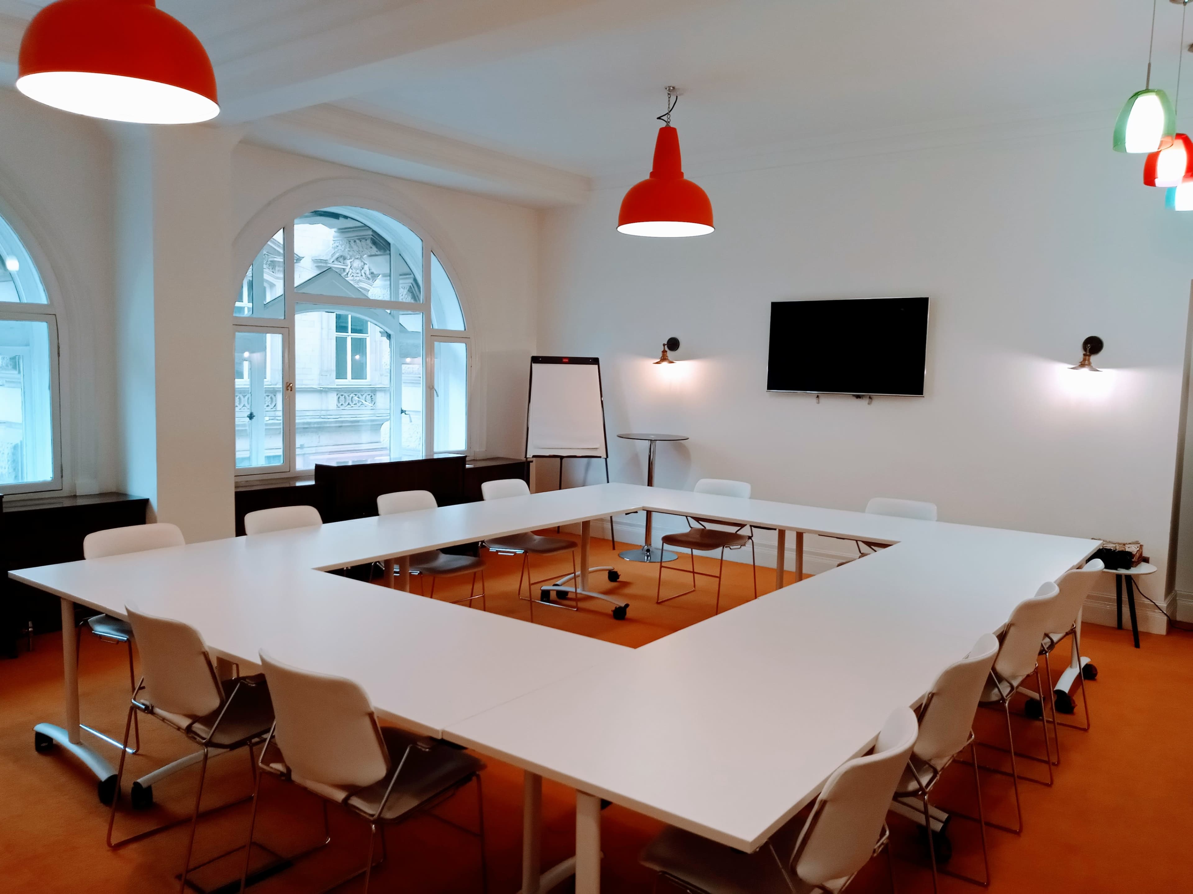 A conference room featuring a large square table surrounded by white chairs, with large windows, a television, and several pendant lights hanging from the ceiling.