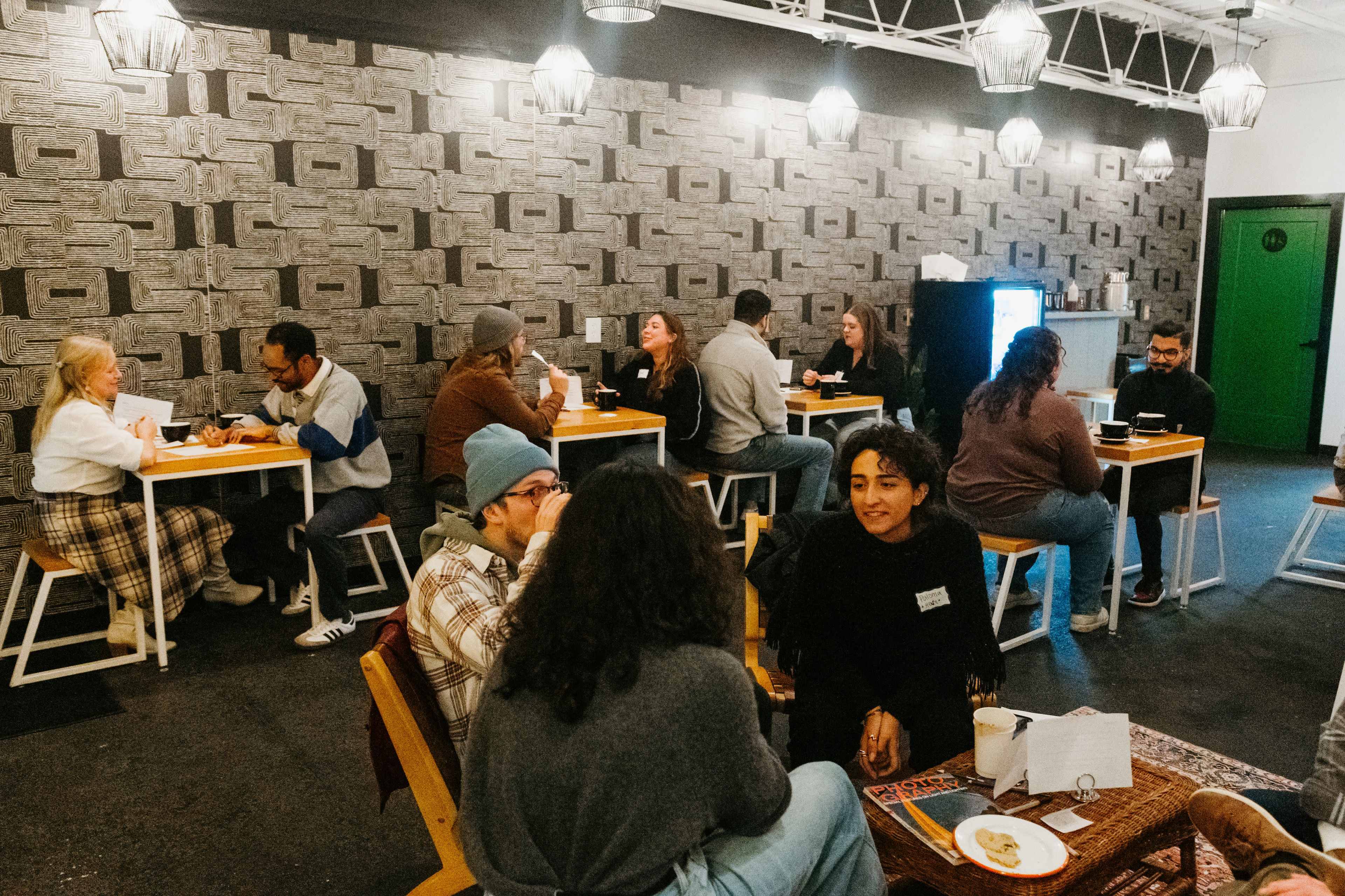 The image shows a lively café interior with groups of people seated at tables, engaged in conversation against a textured wall and hanging light fixtures.