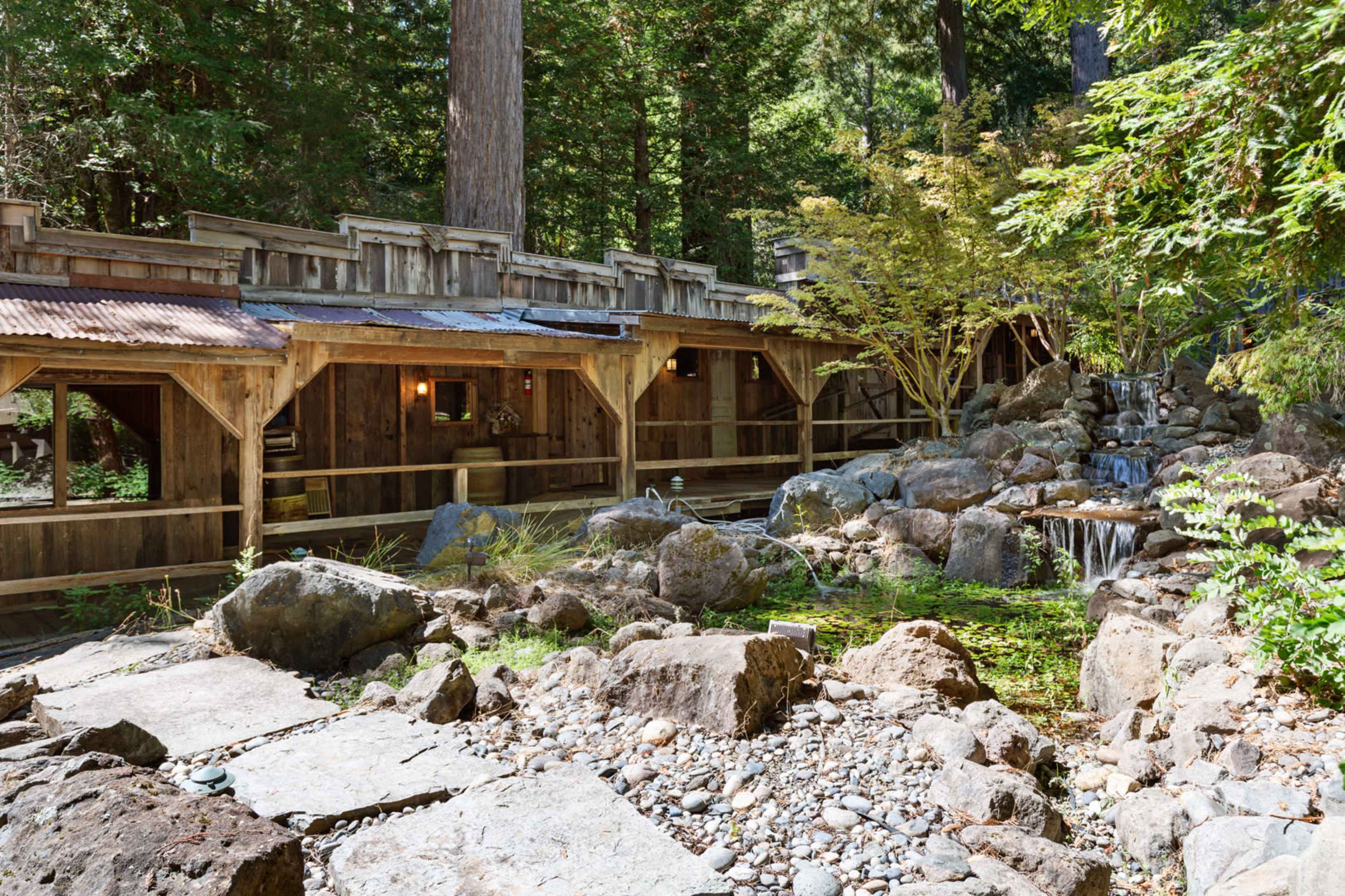 The image shows a rustic wooden structure set among large rocks and greenery, with a small waterfall and pond in the foreground.
