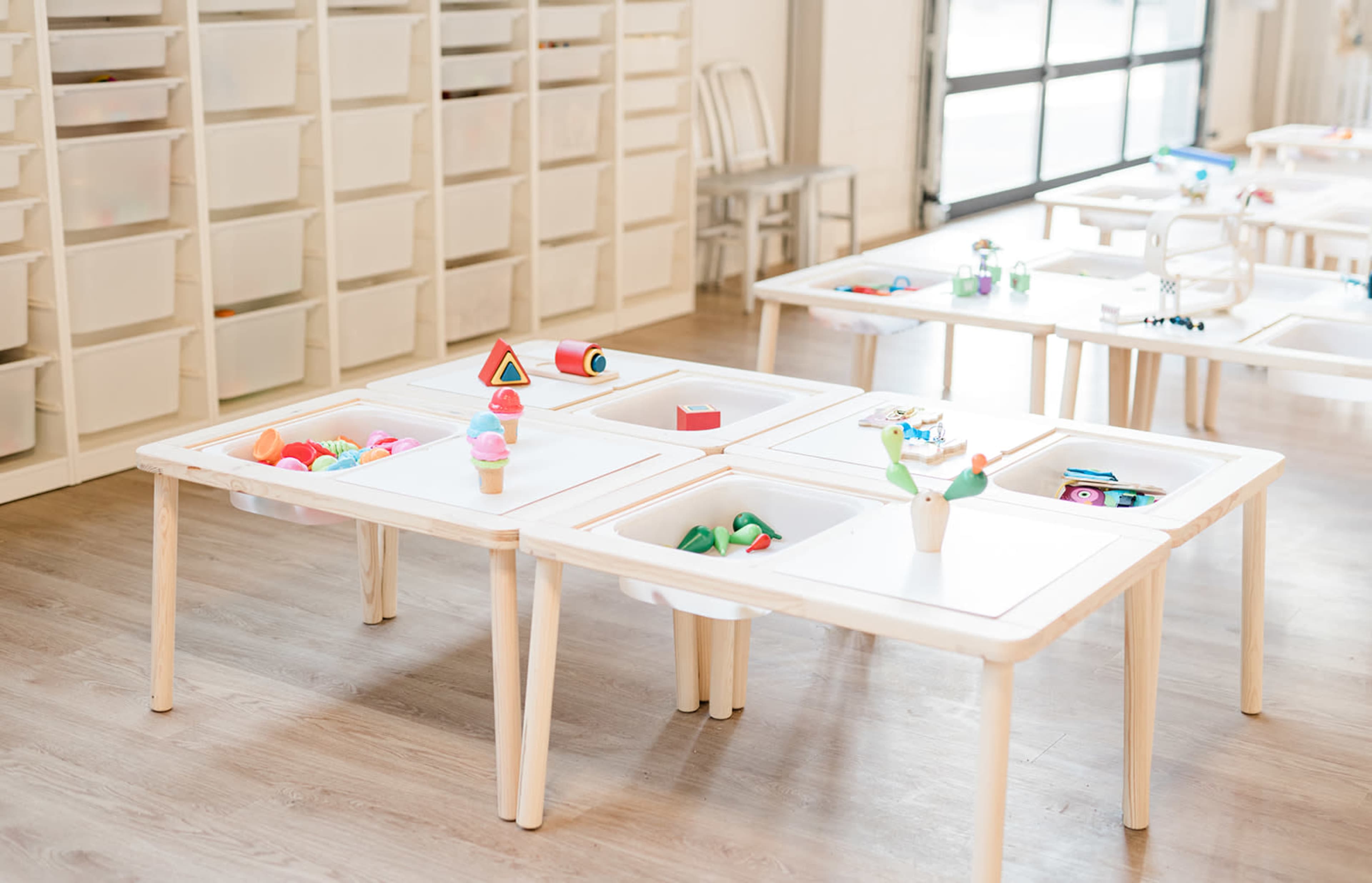 A classroom filled with white tables, each with various colorful educational toys and materials arranged in their center.