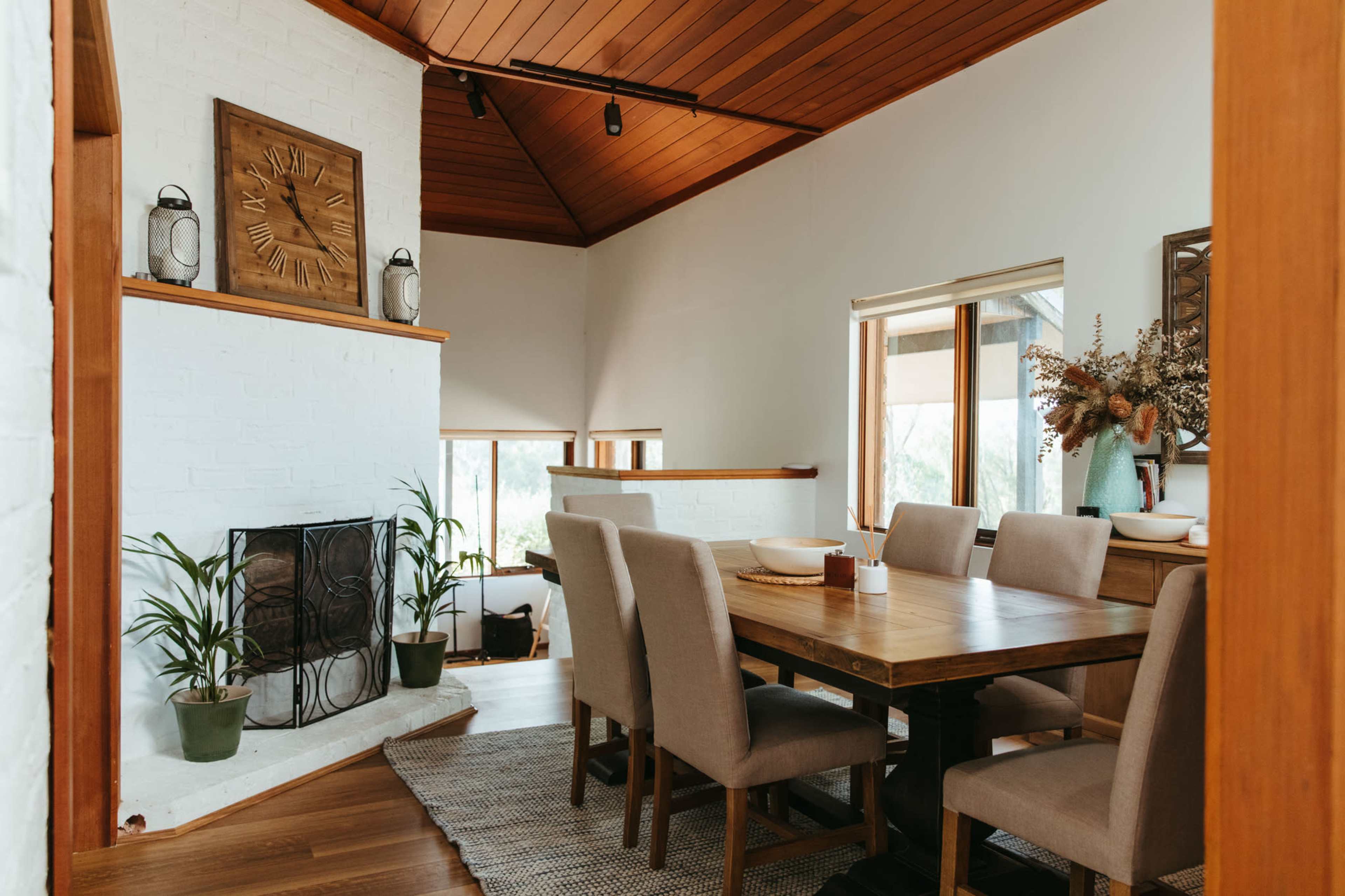 A dining area featuring a large wooden table surrounded by upholstered chairs, with a fireplace and decorative elements on the walls.