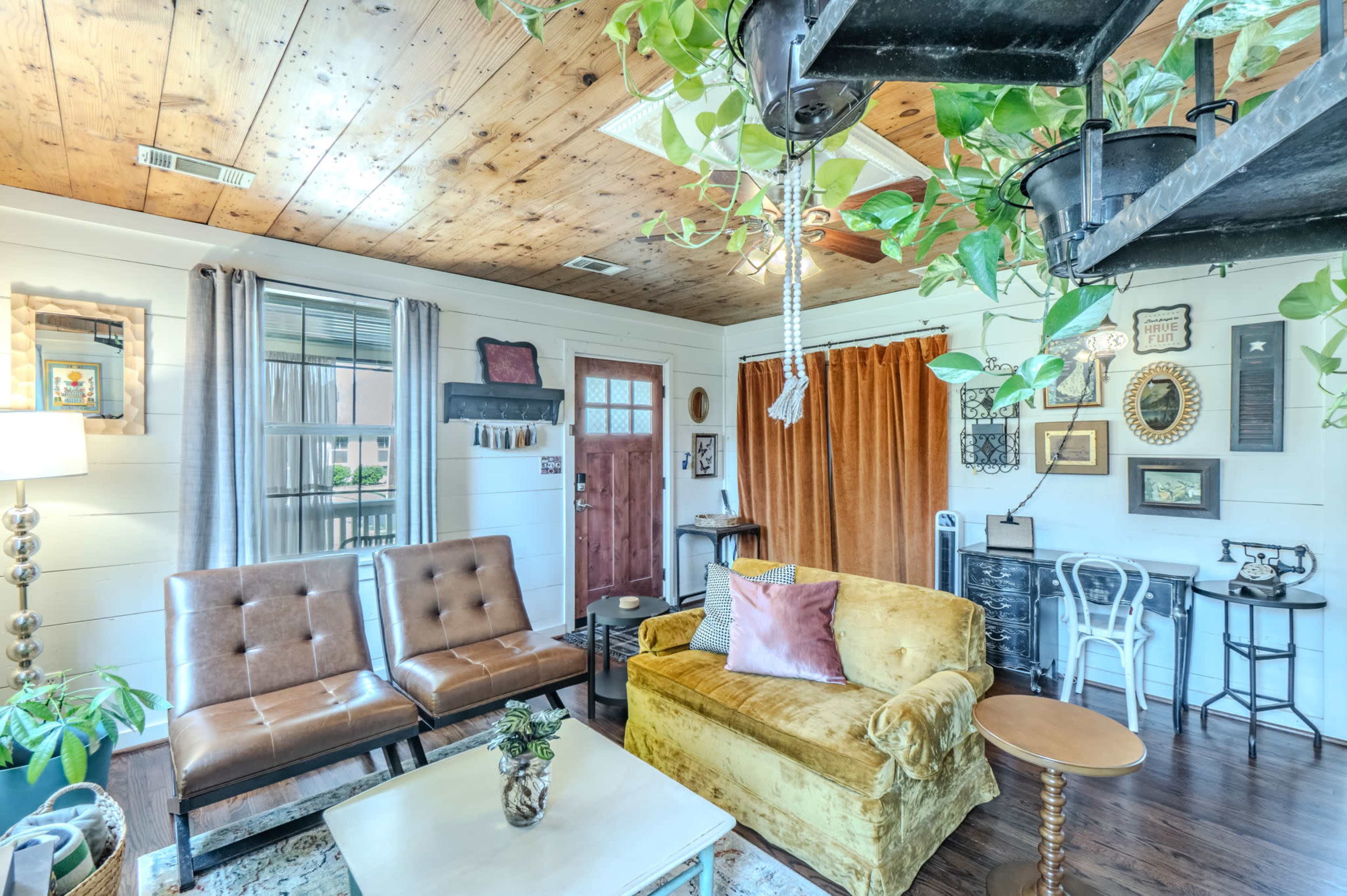 The image shows a cozy living room with a mix of seating options, including a yellow sofa and brown chairs, along with decorative plants and framed pictures on the walls.