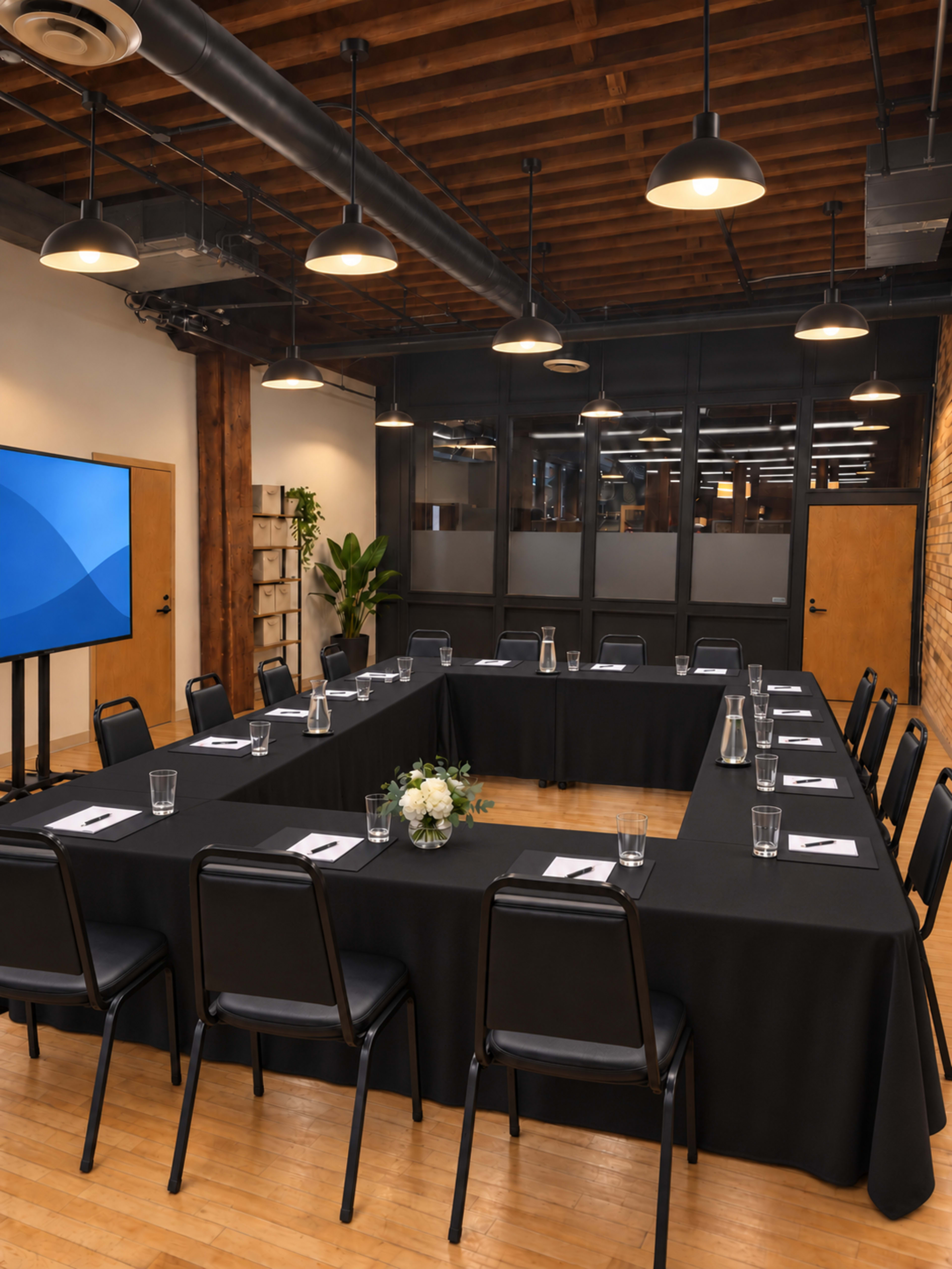 The image shows a rectangular conference room with a long table set up for a meeting, surrounded by black chairs and illuminated by overhead lights.