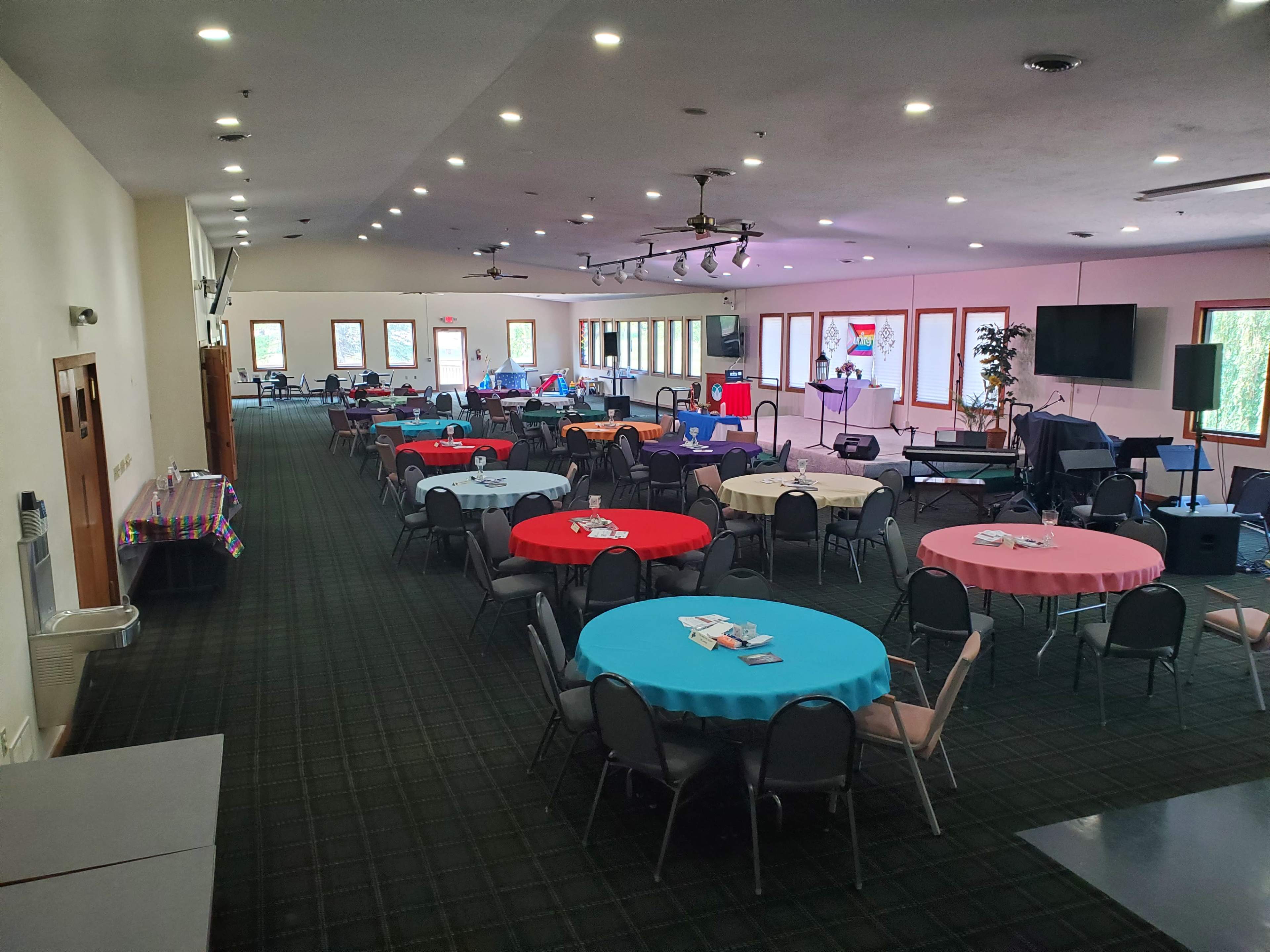 The image shows a spacious event hall with several round tables covered in colorful tablecloths, arranged in a well-lit setting with a stage and equipment at one end.