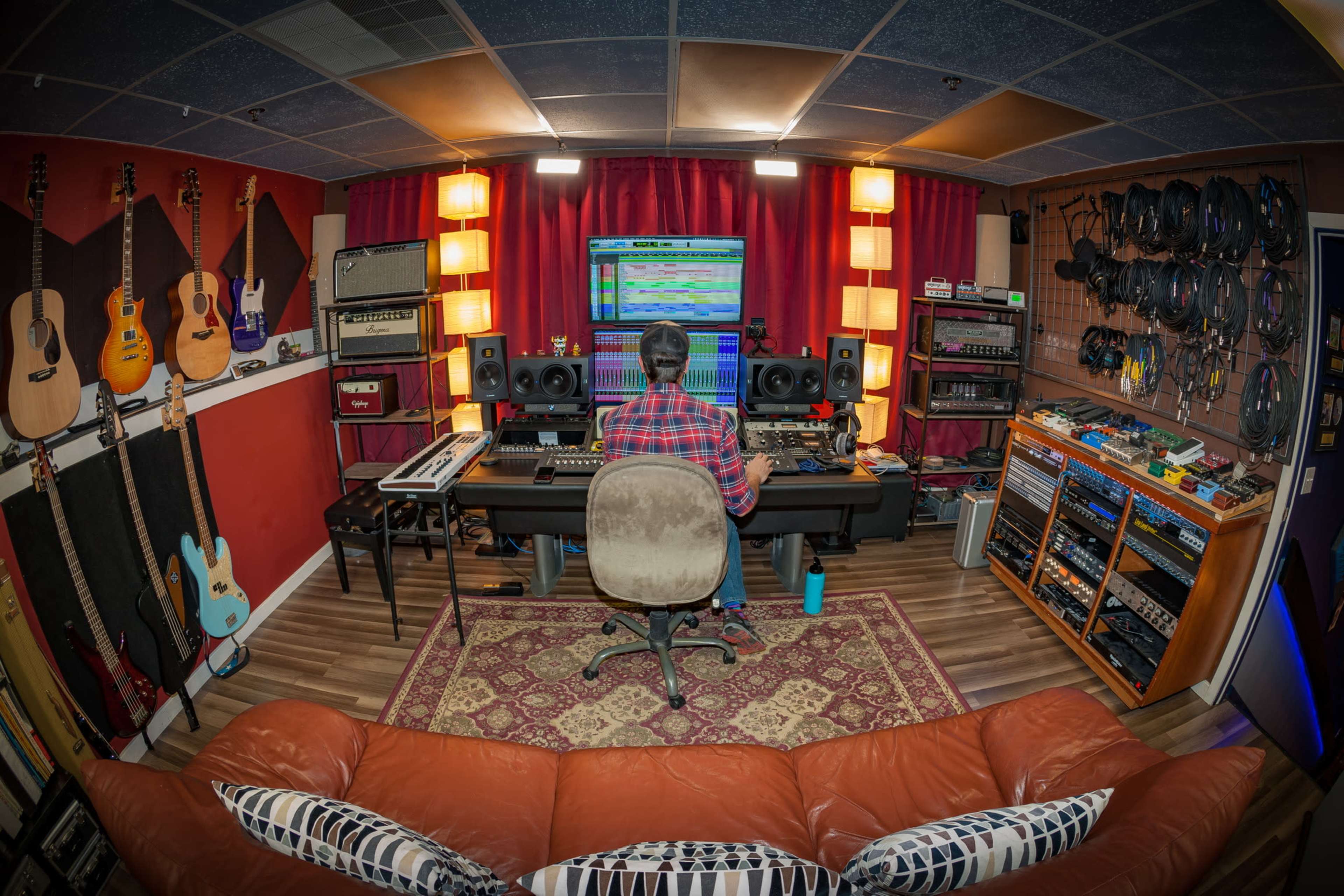 A person sits at a computer workstation in a music studio surrounded by various musical instruments and equipment.