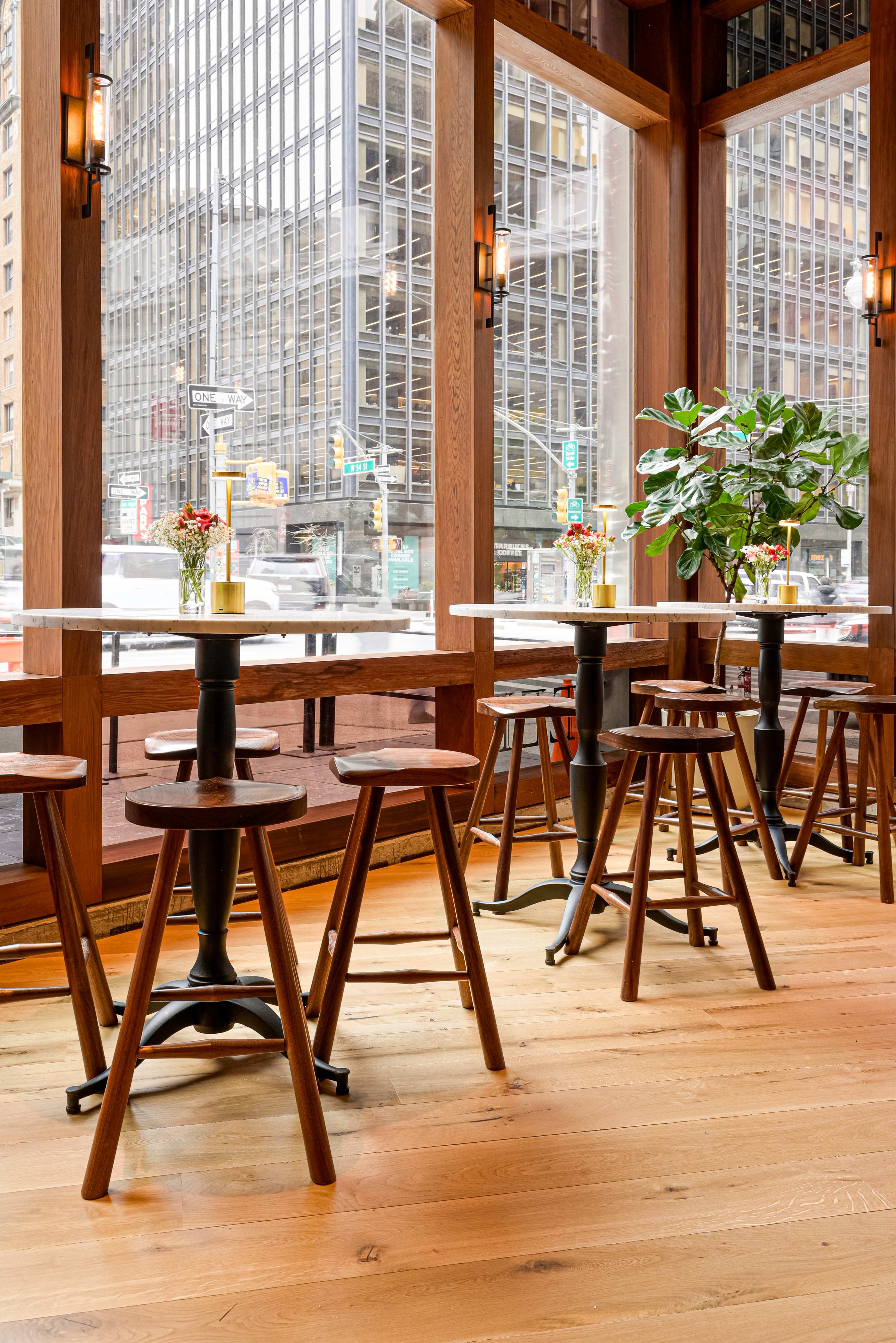 The image shows a modern cafe interior featuring wooden stools and tables near large windows with city views.